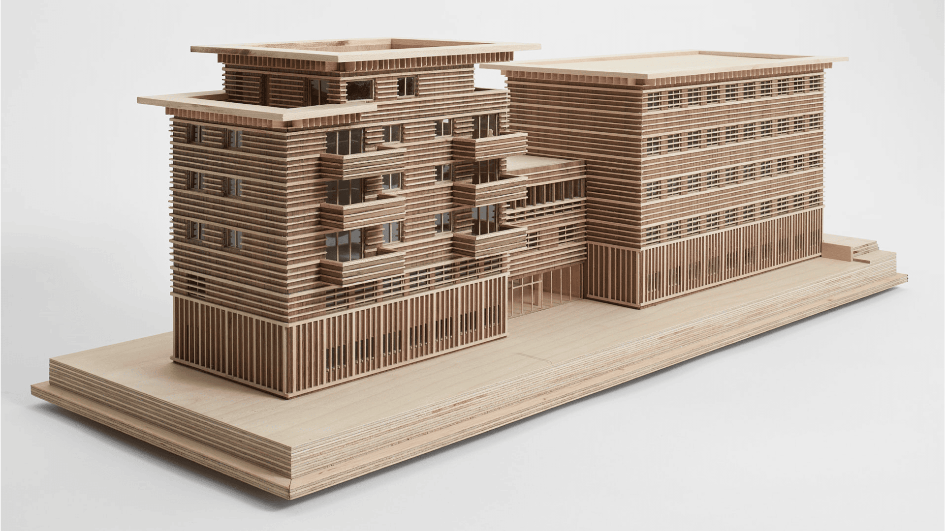 Ein Architekturmodell aus Holz zeigt ein modernes mehrstöckiges Gebäude mit horizontalen Lamellen und großen Fenstern auf einem rechteckigen Sockel vor einem schlichten weißen Hintergrund.
