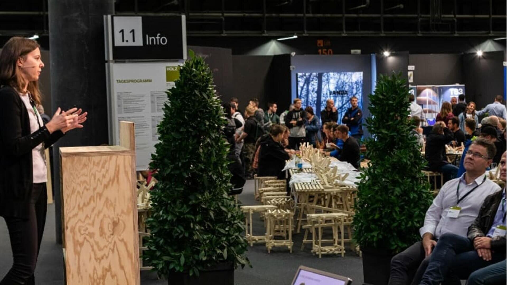 Une femme se tient debout et parle sur un podium devant un public assis parmi des plantes et des chaises en bois lors d'une conférence ou d'un événement en intérieur. Un panneau d'information et des stands d'exposition sont visibles à l'arrière-plan.