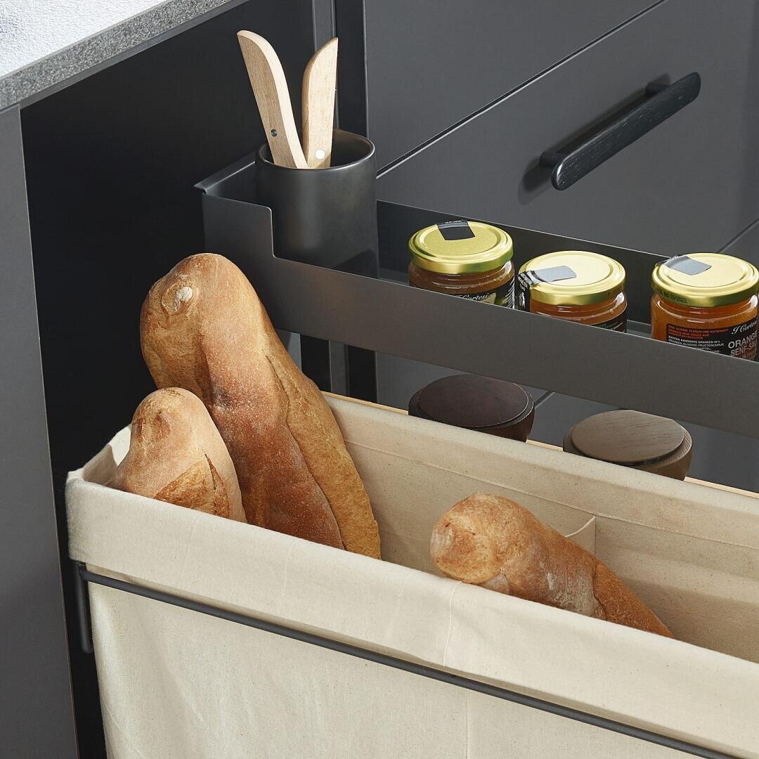 Eine Küchenschublade mit zwei Baguettes in einem beigen Stoffbehälter und Gläsern mit Konserven oder Aufstrichen auf einem Regal, daneben Holzutensilien in einem schwarzen Behälter.