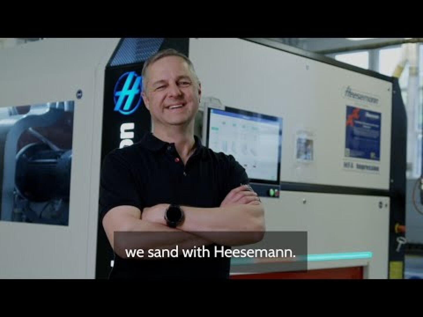 Une personne souriante se tient les bras croisés devant une grande ponceuse industrielle Heesemann. Le sous-titre est le suivant : "we sand with Heesemann".