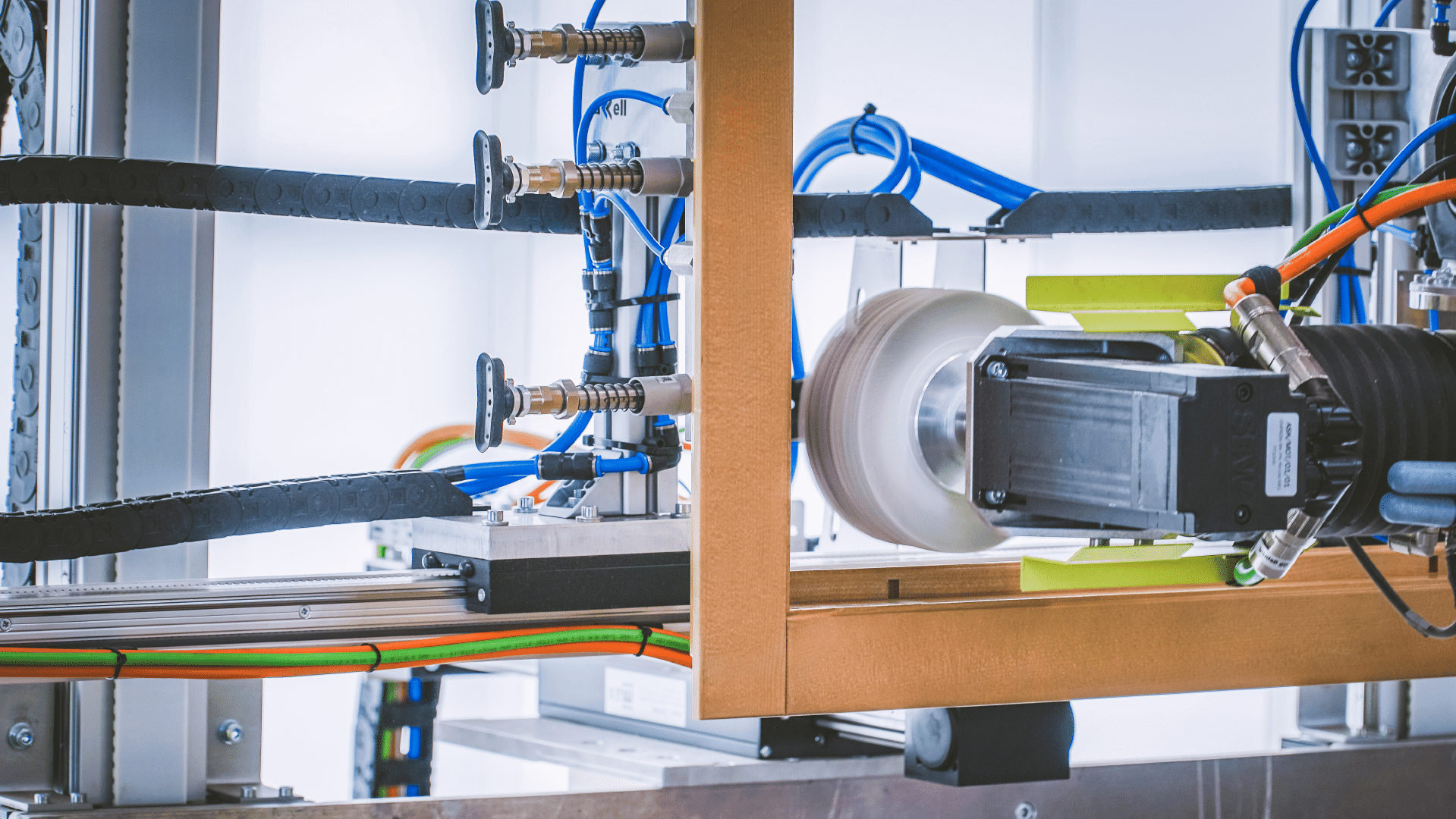 Nahaufnahme einer Industriemaschine mit Kabeln, Rohren und einer motorisierten Komponente mit einem weißen, runden Teil, wahrscheinlich Teil einer automatisierten Fertigungs- oder Montagelinie.