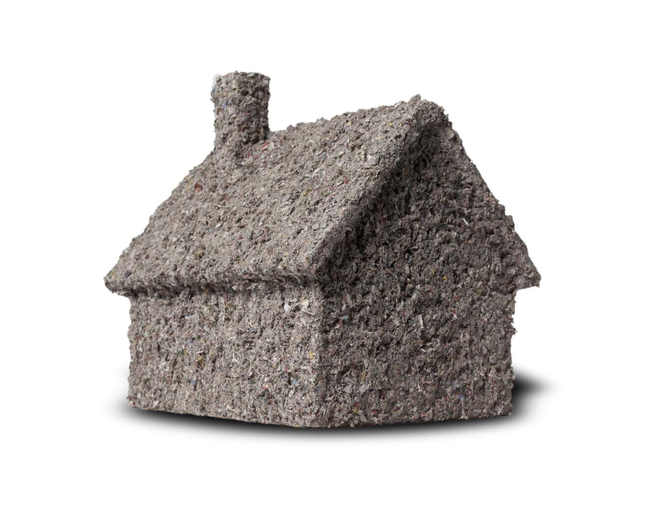Ein kleines Hausmodell aus grauem, rauem, faserigem Material, das an recyceltes Papier oder Dämmstoff erinnert, mit einem Schornstein, der vor einem weißen Hintergrund steht und einen weichen Schatten wirft.