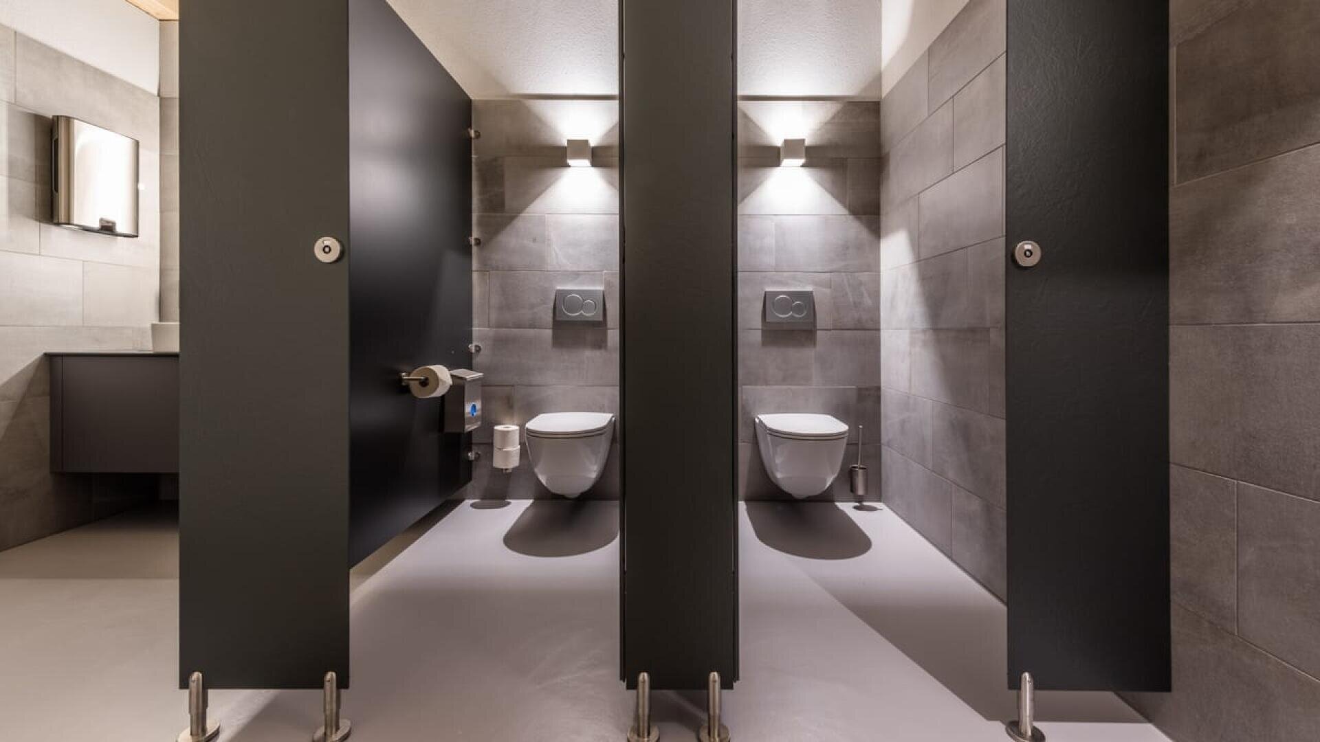 Trois cabines de toilettes modernes avec des cloisons grises et des murs carrelés, chacune contenant des toilettes murales, un porte-papier hygiénique et une petite poubelle. Les cabines sont bien éclairées et propres.