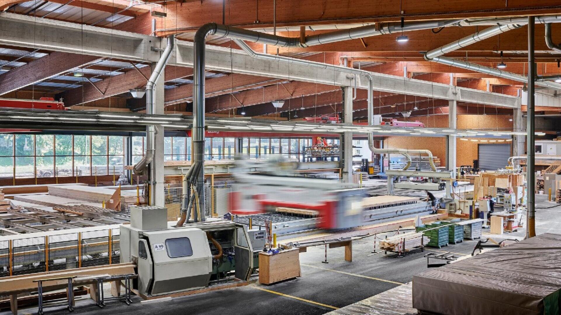 Un intérieur d'usine spacieux et moderne avec de grandes machines à travailler le bois, des piles de bois et des éclairages aériens. La lumière du soleil passe à travers de grandes fenêtres, éclairant les zones de travail et l'équipement.