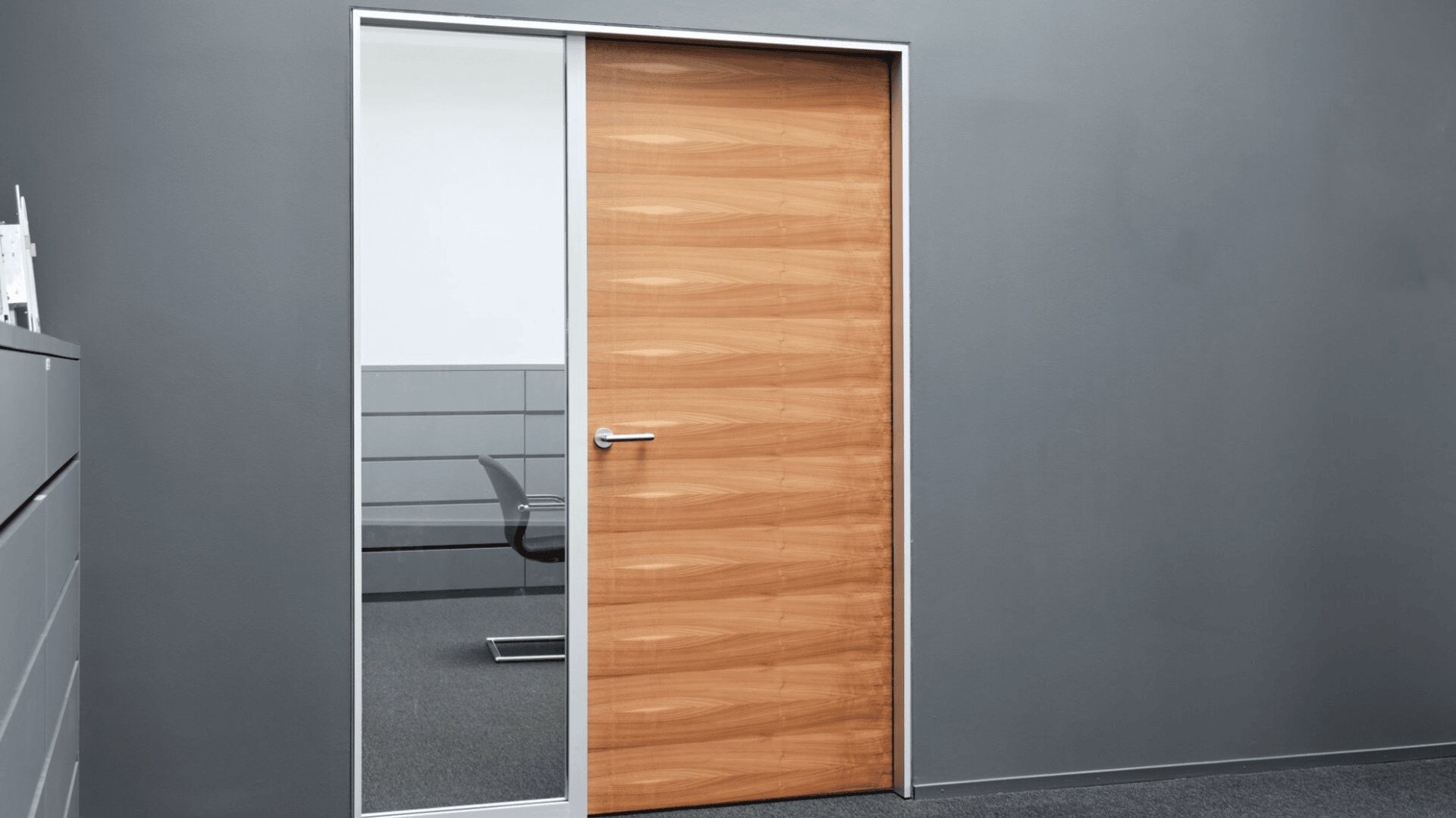 Un intérieur de bureau moderne avec une porte en bois fermée et une grande vitre à côté qui montre une partie d'une chaise et d'un bureau dans une pièce adjacente. Les murs sont gris et le sol est recouvert de moquette.
