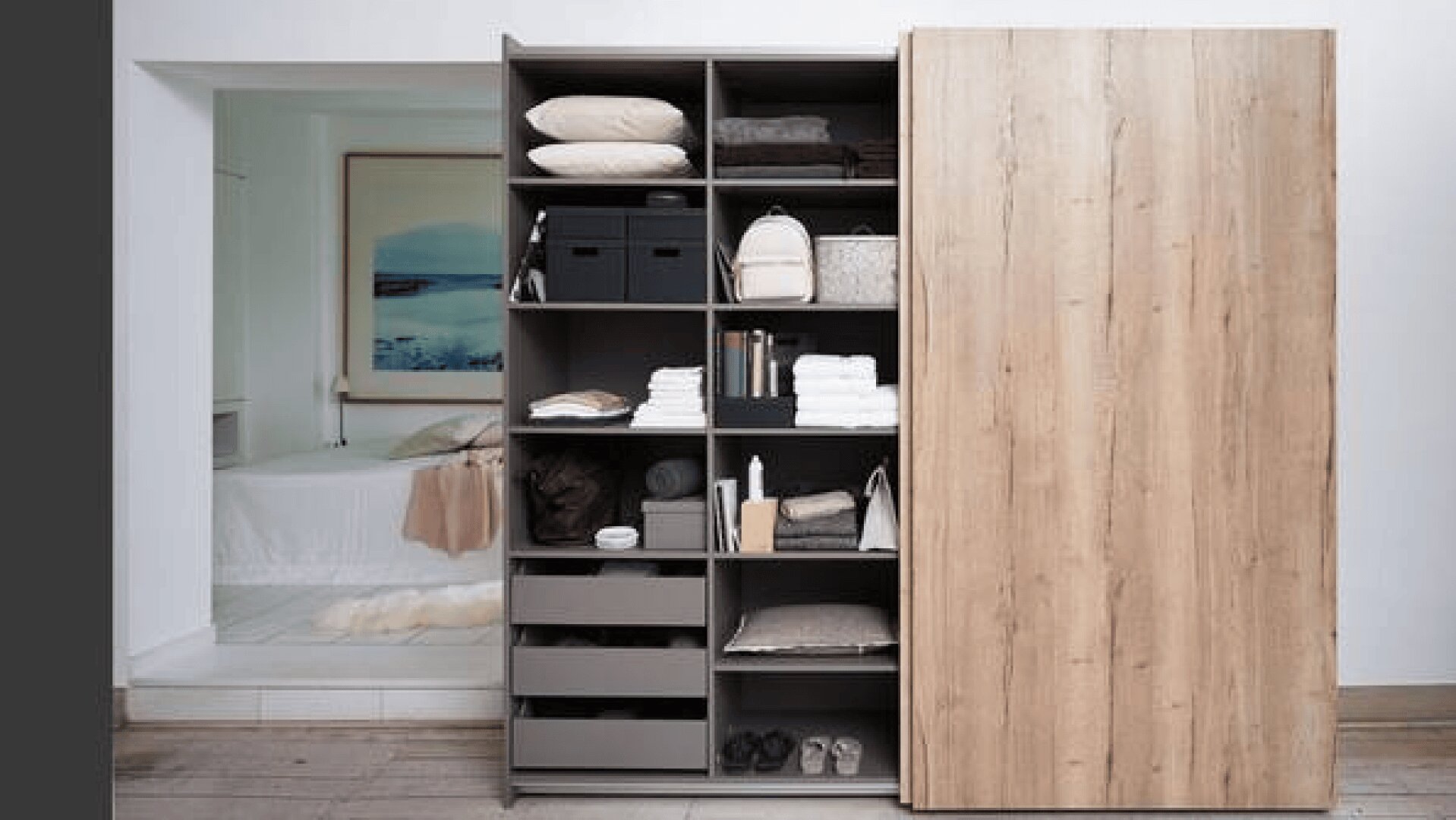 Offener moderner Kleiderschrank mit Regalen, die mit ordentlich organisierter Kleidung, Handtüchern, Taschen, Kisten und Kissen gefüllt sind. Die Schiebetür aus Holz verdeckt teilweise die rechte Seite. Schlafzimmer mit Bett im Hintergrund sichtbar.