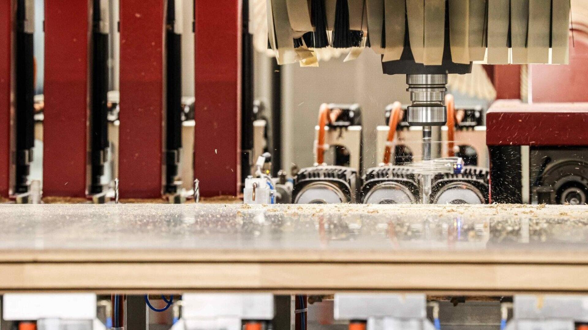 Vue rapprochée d'une machine CNC coupant et façonnant une pièce de bois, avec de la sciure et des copeaux de bois visibles sur la surface de travail. Des machines automatisées et des composants sont visibles à l'arrière-plan.