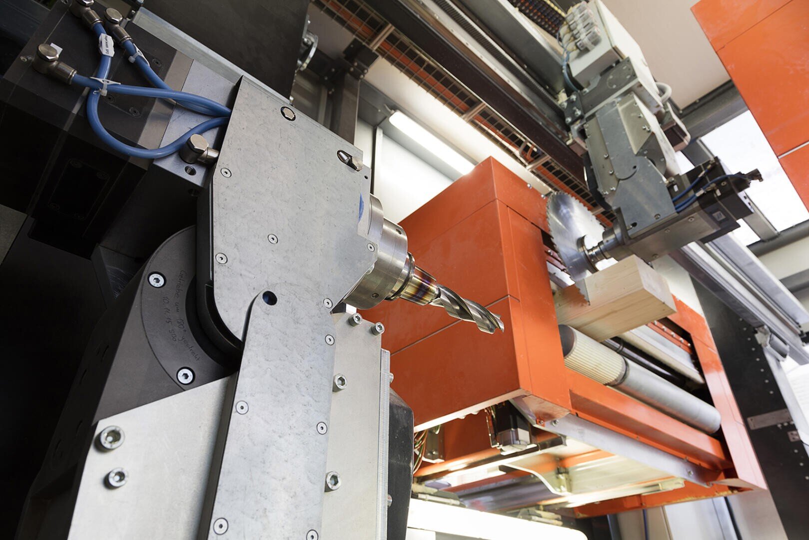 Nahaufnahme einer industriellen CNC-Maschine mit einem Bohrer, Metallkomponenten und einem orangefarbenen Gehäuse, die in einer modernen Produktionsumgebung auf einem Holzblock arbeitet.