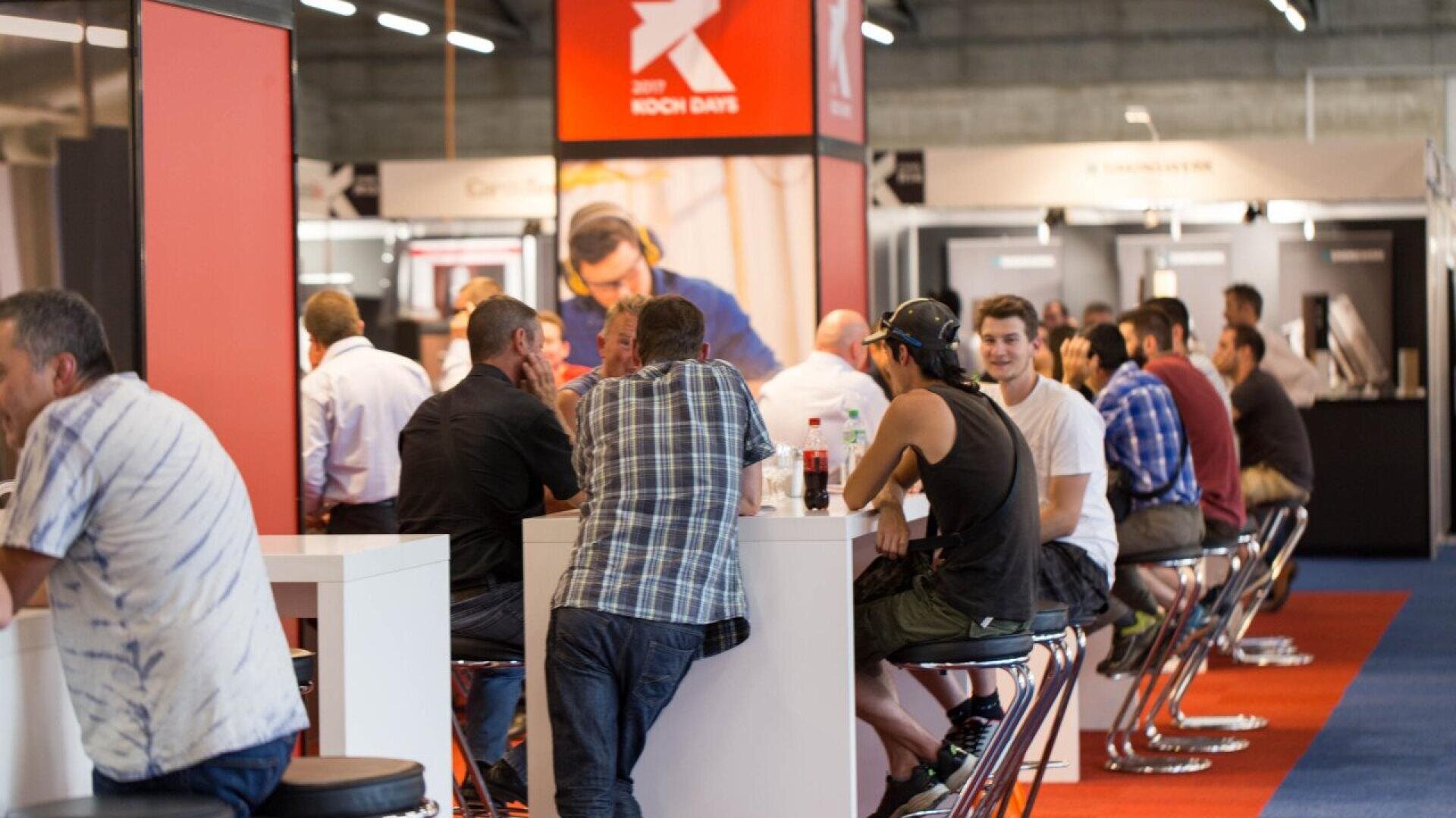 Des personnes sont assises et discutent autour de grandes tables lors d'un événement ou d'une exposition en intérieur très fréquenté, avec des stands d'exposition et des étalages visibles en arrière-plan. L'atmosphère est décontractée et sociale, avec un mélange d'hommes qui discutent et se détendent.