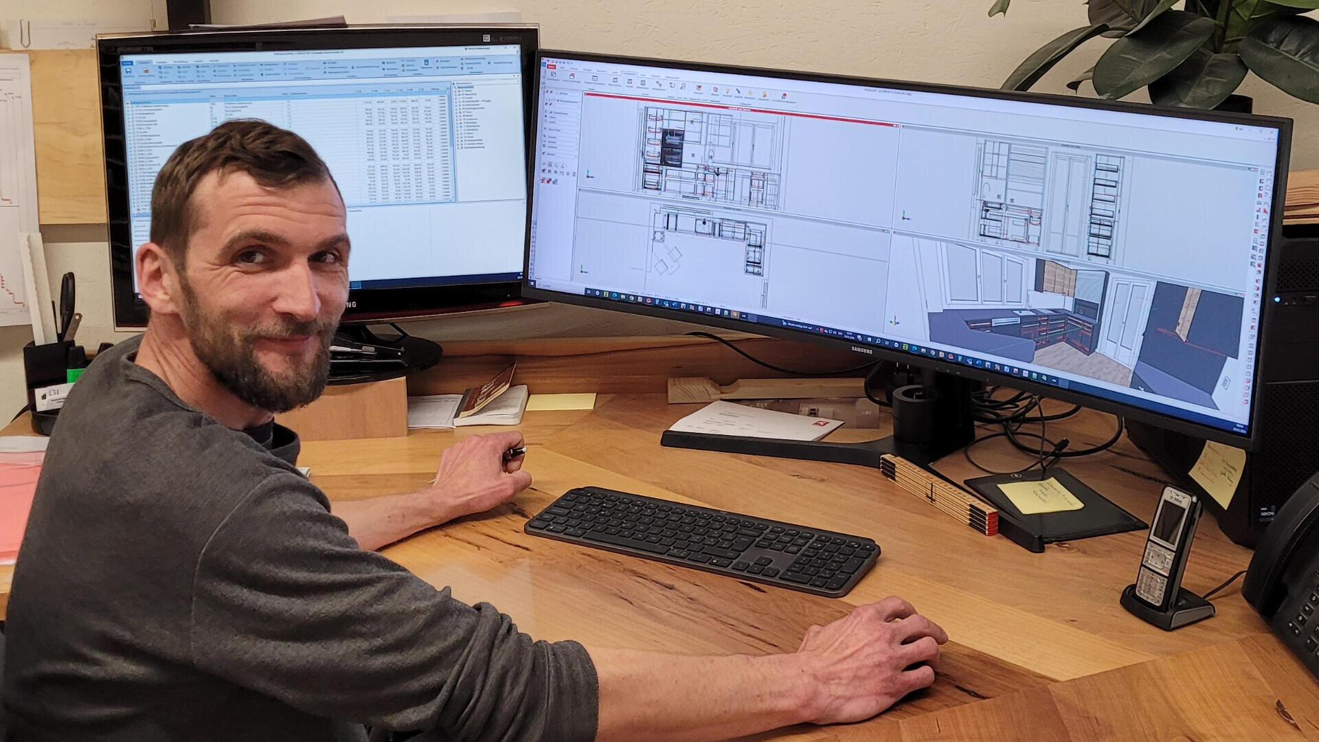 Ein Mann sitzt an einem hölzernen Schreibtisch und lächelt in die Kamera, während er an einem Computer mit zwei großen Monitoren arbeitet, auf denen Architektur- oder Innenarchitektur-Software und detaillierte Raumpläne angezeigt werden.