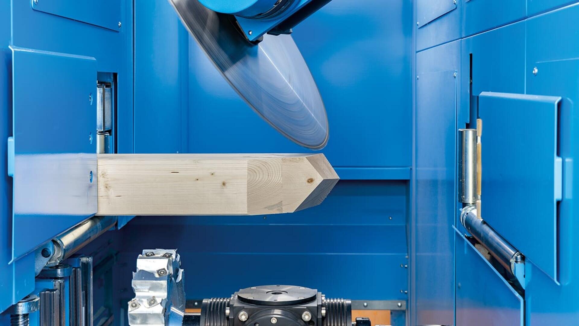 Ein Kreissägeblatt in einer blauen Industriemaschine schneidet das Ende eines Holzbalkens in einem Winkel, wobei Maschinen und Rollen um das Holz herum sichtbar sind.