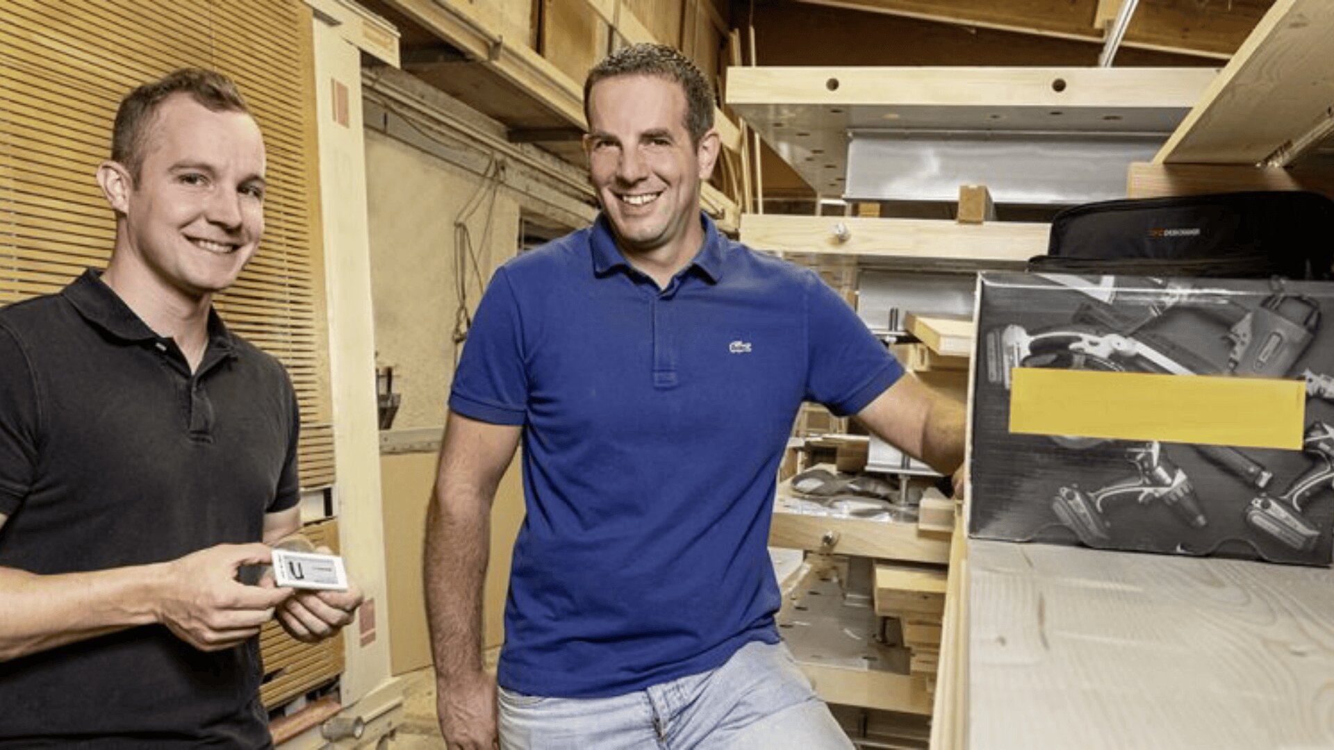 Zwei Männer stehen lächelnd in einer Werkstatt mit Holzregalen und Eisenwarenwerkzeugen. Einer hält einen kleinen Gegenstand, während der andere sich auf ein Regal stützt. Die Umgebung sieht organisiert und gut beleuchtet aus.