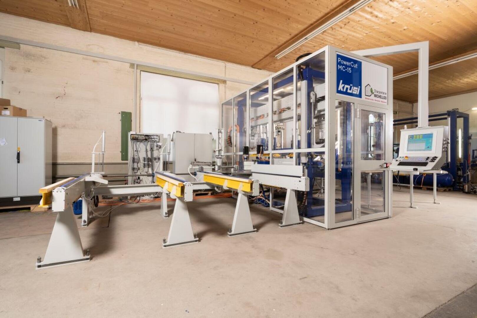 Eine moderne industrielle Holzbearbeitungsmaschine mit Förderbändern, Sicherheitsschranken und einem digitalen Bedienfeld in einer geräumigen Werkstatt mit Holzdecke und Betonwänden.