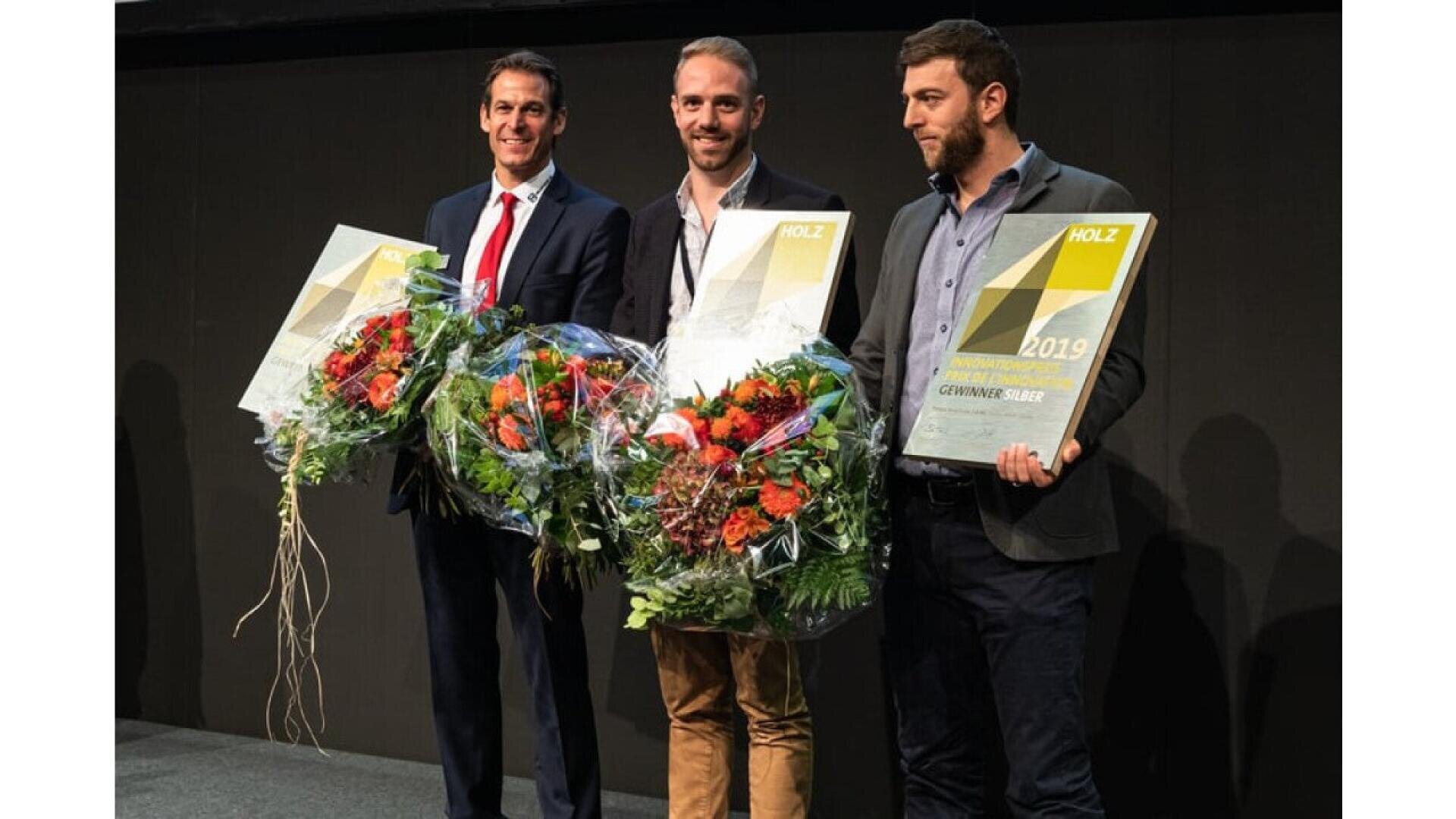 Trois hommes se tiennent côte à côte, tenant de grands bouquets de fleurs et des certificats encadrés, posant pour une photo lors d'une cérémonie de remise de prix. Ils posent pour une photo lors d'une cérémonie de remise de prix. Ils sont tous habillés en tenue de ville ou d'affaires et sourient.