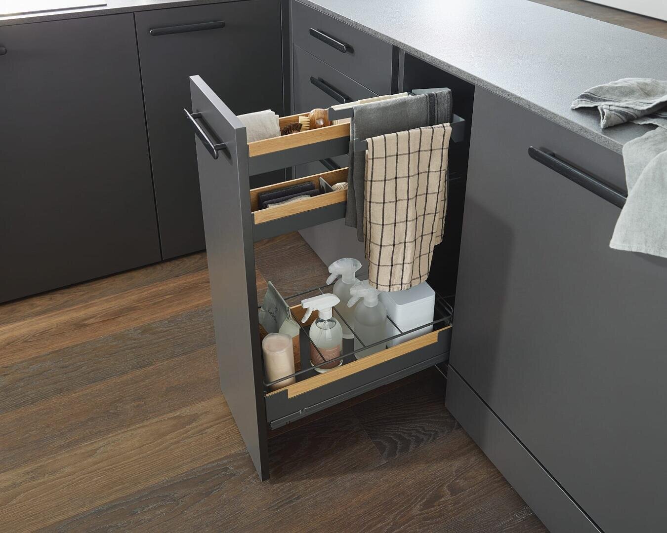 Ein schmaler, ausziehbarer Küchenschrank mit Holzregalen für Reinigungsmittel, Handtücher und Sprühflaschen, der zwischen dunkelgrauen Schränken auf einem braunen Holzboden steht.