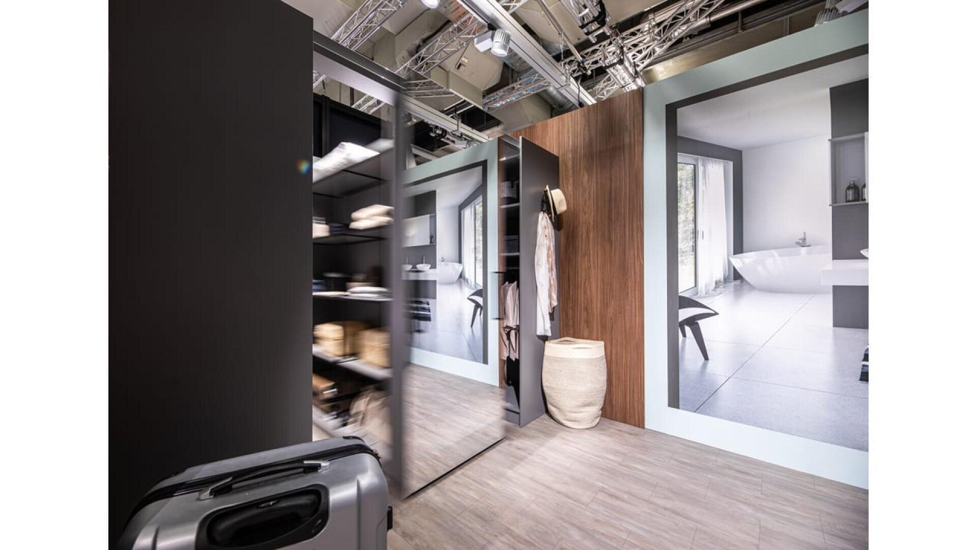 Ein moderner, offener Kleiderschrank mit Regalen und hängenden Kleidungsstücken, mit einem großen Spiegel, einem Weidenkorb und hölzernen Akzenten. Große Wanddisplays zeigen eine minimalistische Badezimmerszene. Ein Koffer steht im Vordergrund.