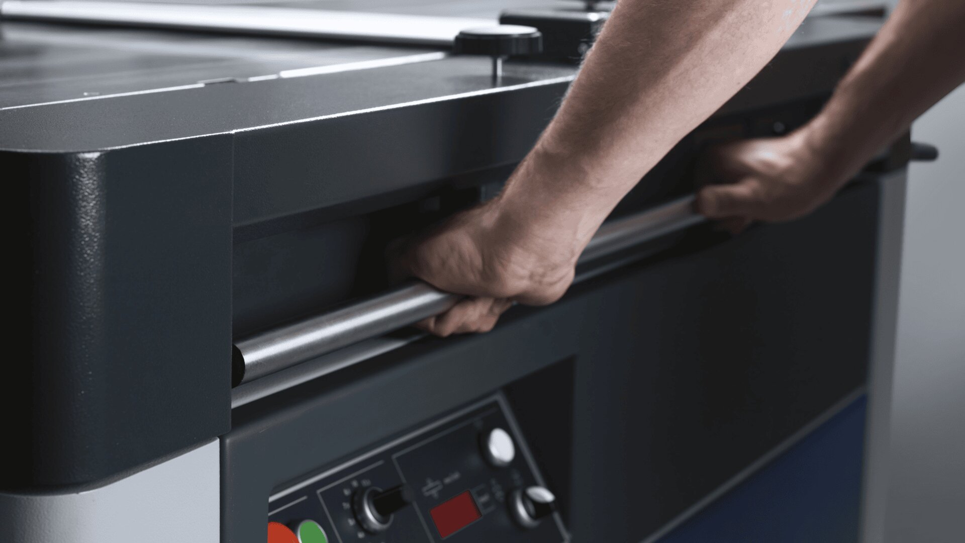Nahaufnahme einer Person, die einen Metallgriff an einer Industriemaschine ergreift. Auf der Vorderseite der Maschine sind Bedientasten und eine Digitalanzeige zu sehen.