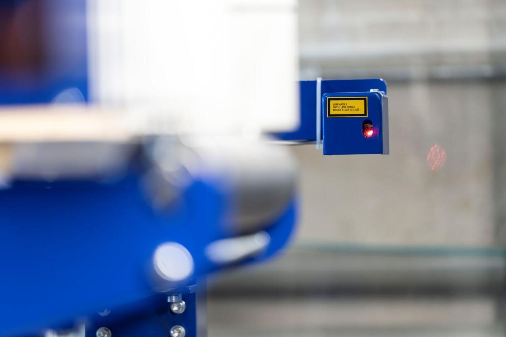 Nahaufnahme eines blauen Industriesensors mit roter Kontrollleuchte, der an einem Metallgerät angebracht ist. Der Sensor ist mit einem gelben Warnhinweis versehen und befindet sich in einer unscharfen Produktions- oder Lagerumgebung.