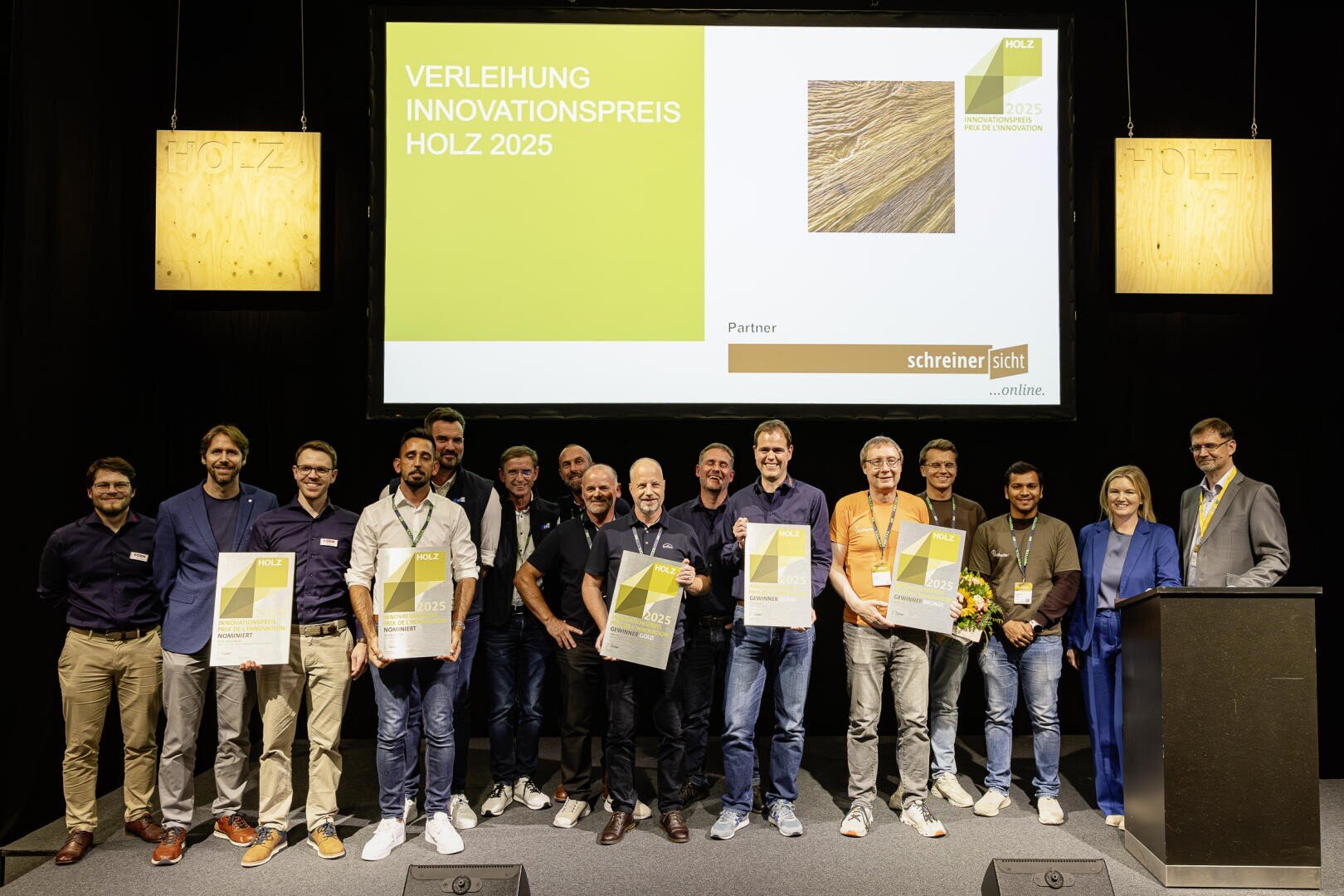 Un groupe de personnes se tient sur la scène et tient des certificats et des fleurs lors de la remise du prix de l'innovation HOLZ 2025. Derrière eux, on peut voir un grand écran de présentation avec les détails de l'événement.