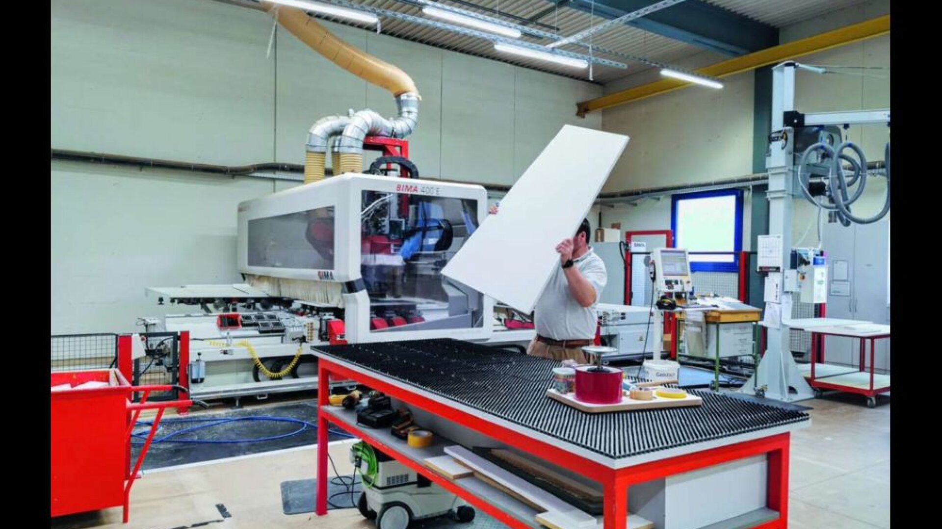 Eine Person bedient Maschinen in einer Fabrikhalle und hält eine große weiße Tafel neben einer industriellen Schneide- oder CNC-Maschine. Auf einer Werkbank in der Nähe sind verschiedene Werkzeuge und Geräte angeordnet.