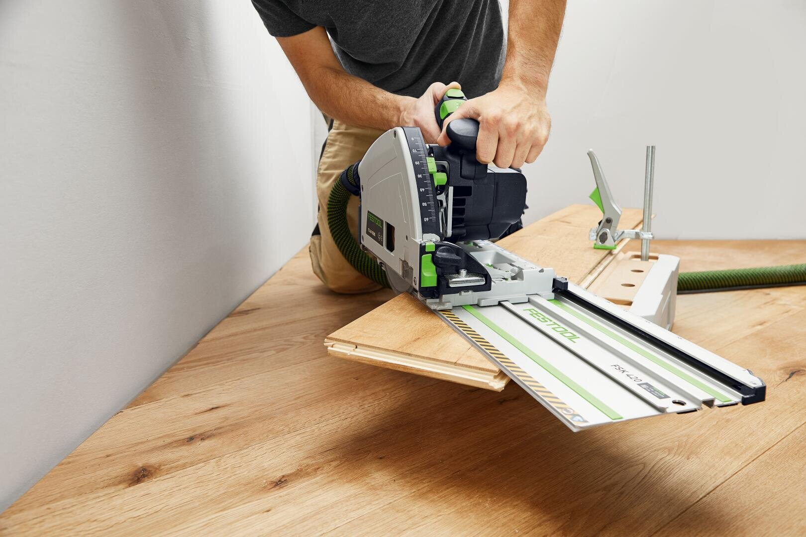 Eine Person sägt mit einer Schienensäge ein Holzbrett auf einer Werkbank, wobei eine Führungsschiene für einen präzisen Schnitt sorgt. Zur Einrichtung gehört ein an der Säge befestigter Staubabsaugschlauch.