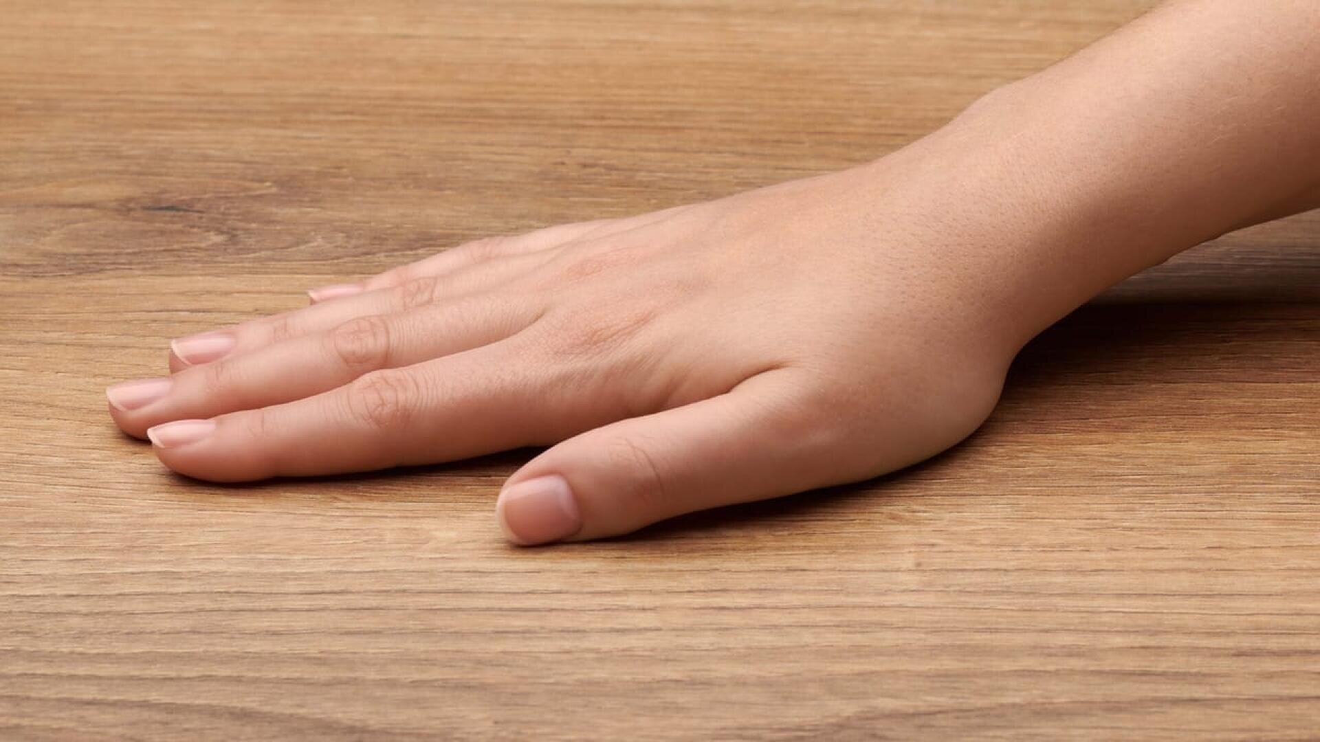 Une main gauche aux ongles naturels et courts repose à plat sur une surface en bois brun clair.