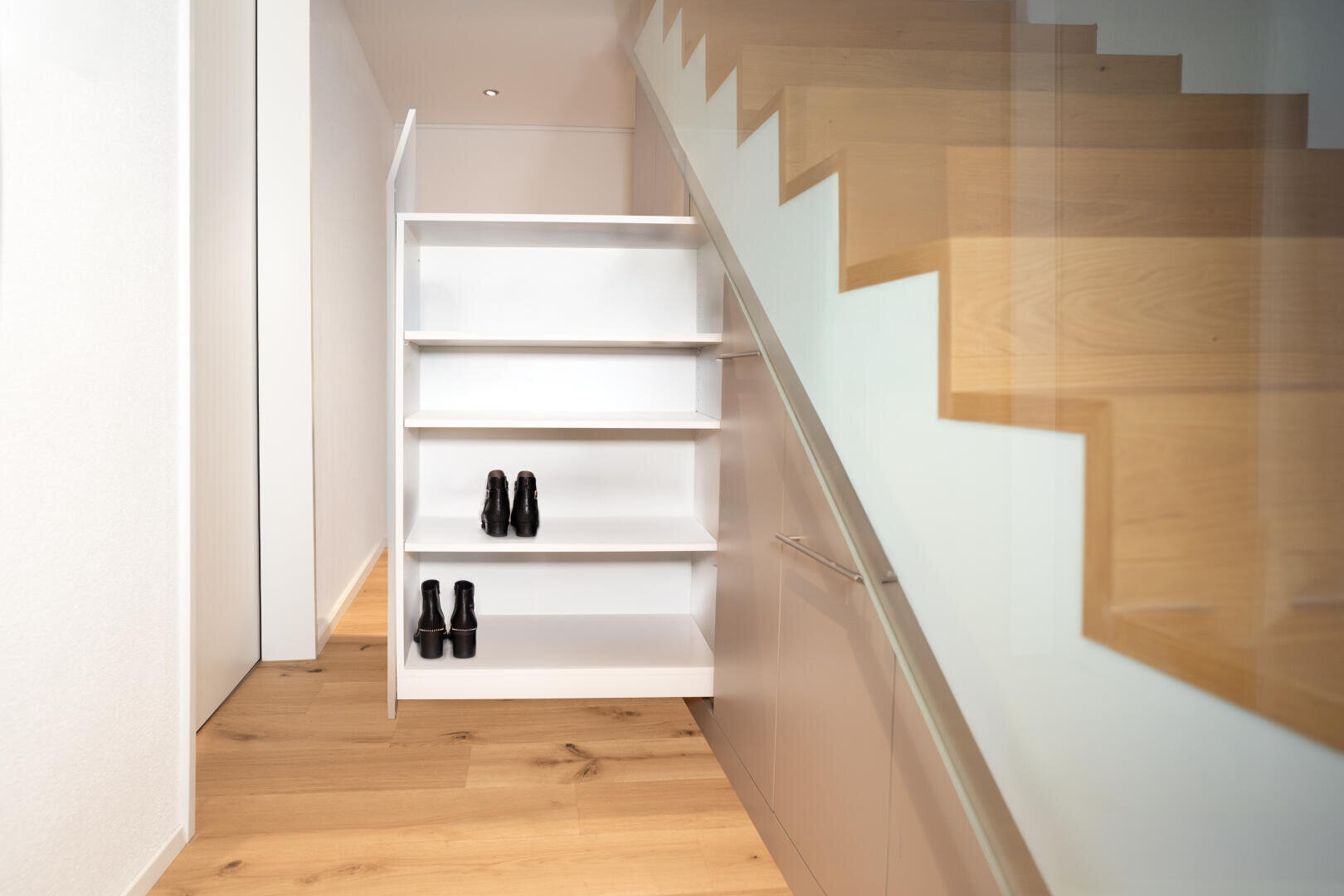 In einem modernen Flur mit Holzfußboden befindet sich unter der Treppe ein ausziehbares weißes Regal, in dem zwei Paar schwarze Schuhe stehen. Die Treppe hat ein Glasgeländer.