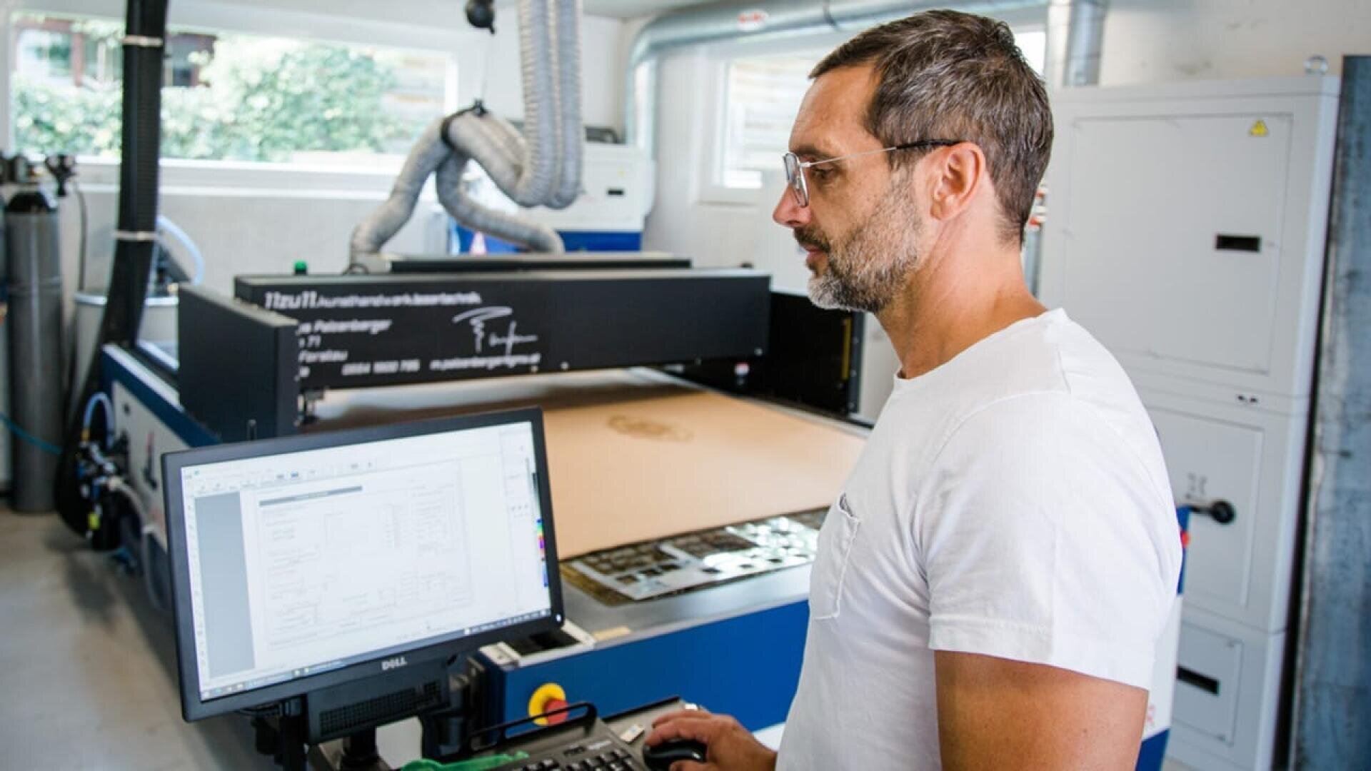 Un homme vêtu d'un t-shirt blanc utilise un ordinateur devant une grande machine industrielle dans un atelier. Il regarde l'écran, des machines et des équipements étant visibles à l'arrière-plan.