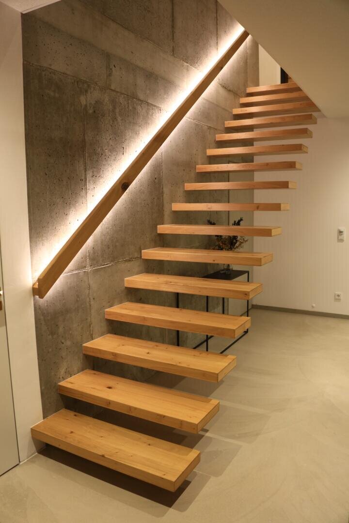 Eine moderne, schwebende Holztreppe mit offenen Setzstufen ist an einer Betonwand befestigt und wird von einer eleganten, diagonalen LED-Leuchte beleuchtet, die ein minimalistisches und zeitgemäßes Innendesign schafft.