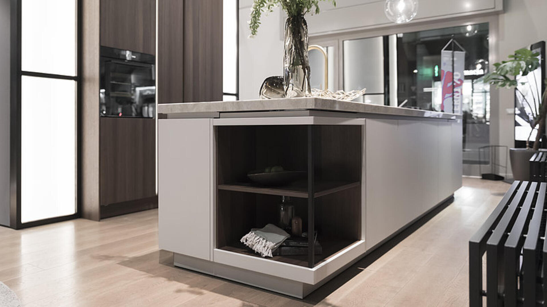 Moderne Küche mit einer großen Insel mit offenen Regalen, dekorativen Gegenständen und einer eingebauten Spüle. Der Raum ist mit Holzböden, minimalistischen Schränken und neutralen Tönen mit sanfter Beleuchtung ausgestattet.