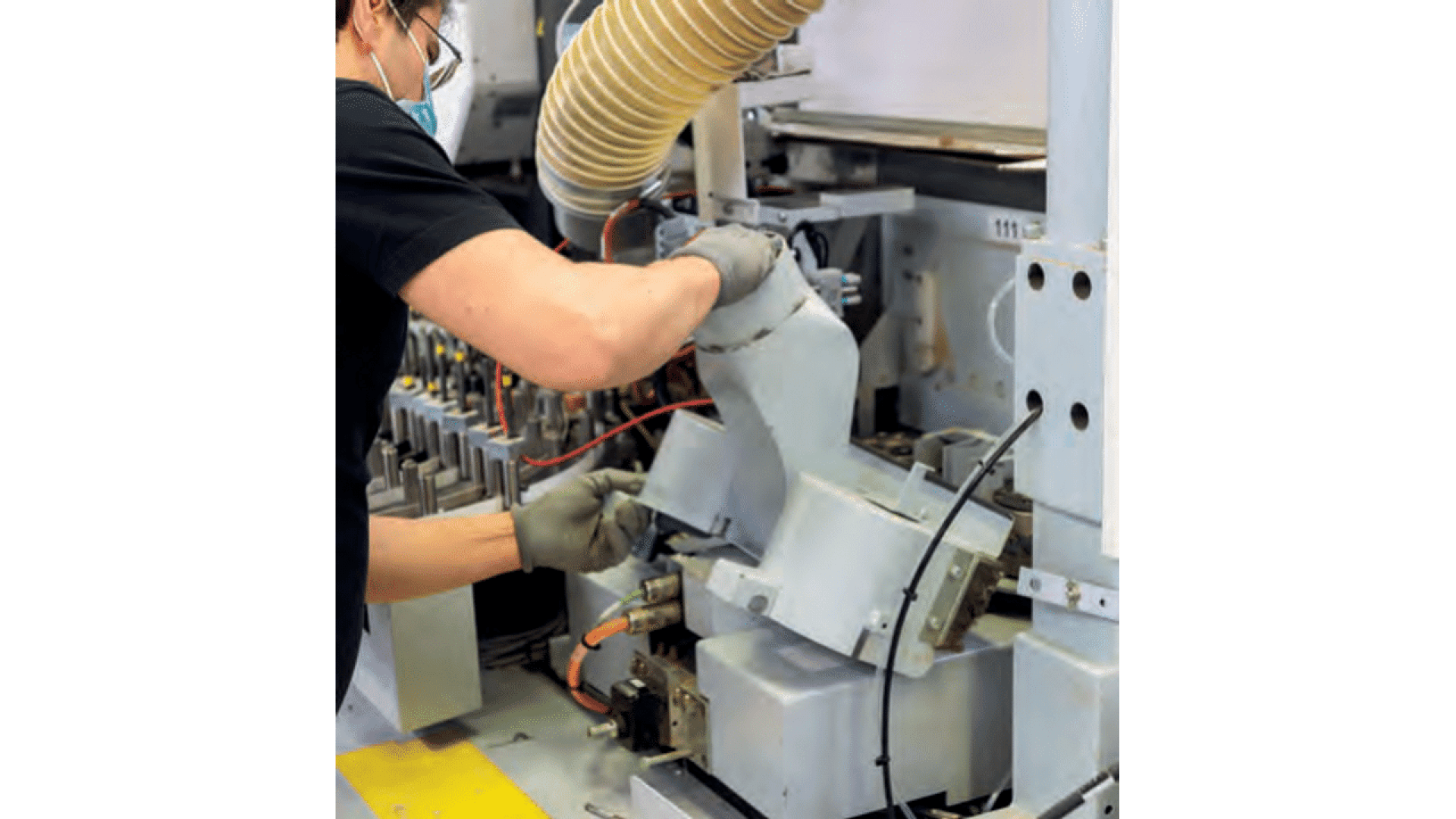 Une personne portant des lunettes de sécurité, un masque et des gants manipule des machines industrielles et tient un gros composant gris sous un tuyau jaune dans une usine.