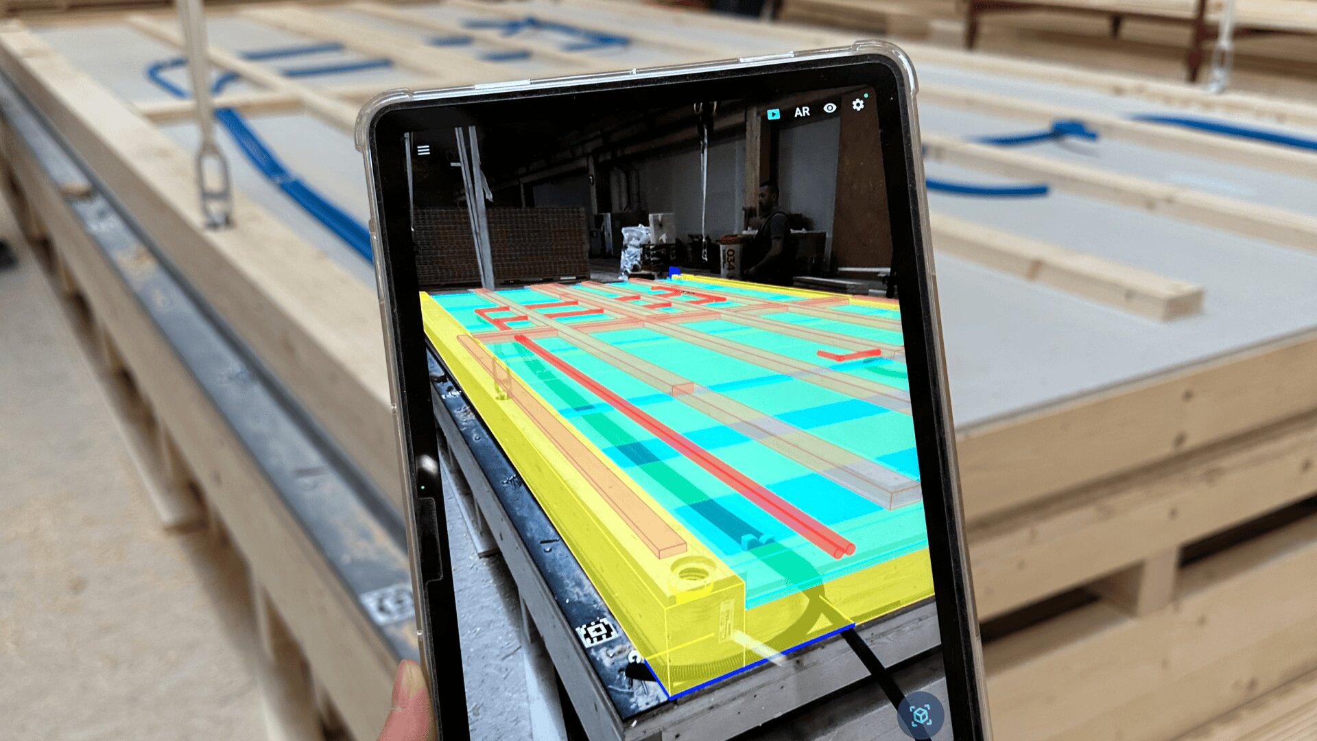 Eine Person hält ein Tablet in der Hand, das ein Augmented-Reality-Overlay von Bauplänen auf einer Holzkonstruktion in einer Werkstatt zeigt, wobei digitale Linien und Farben mit physischen Materialien abgeglichen werden.