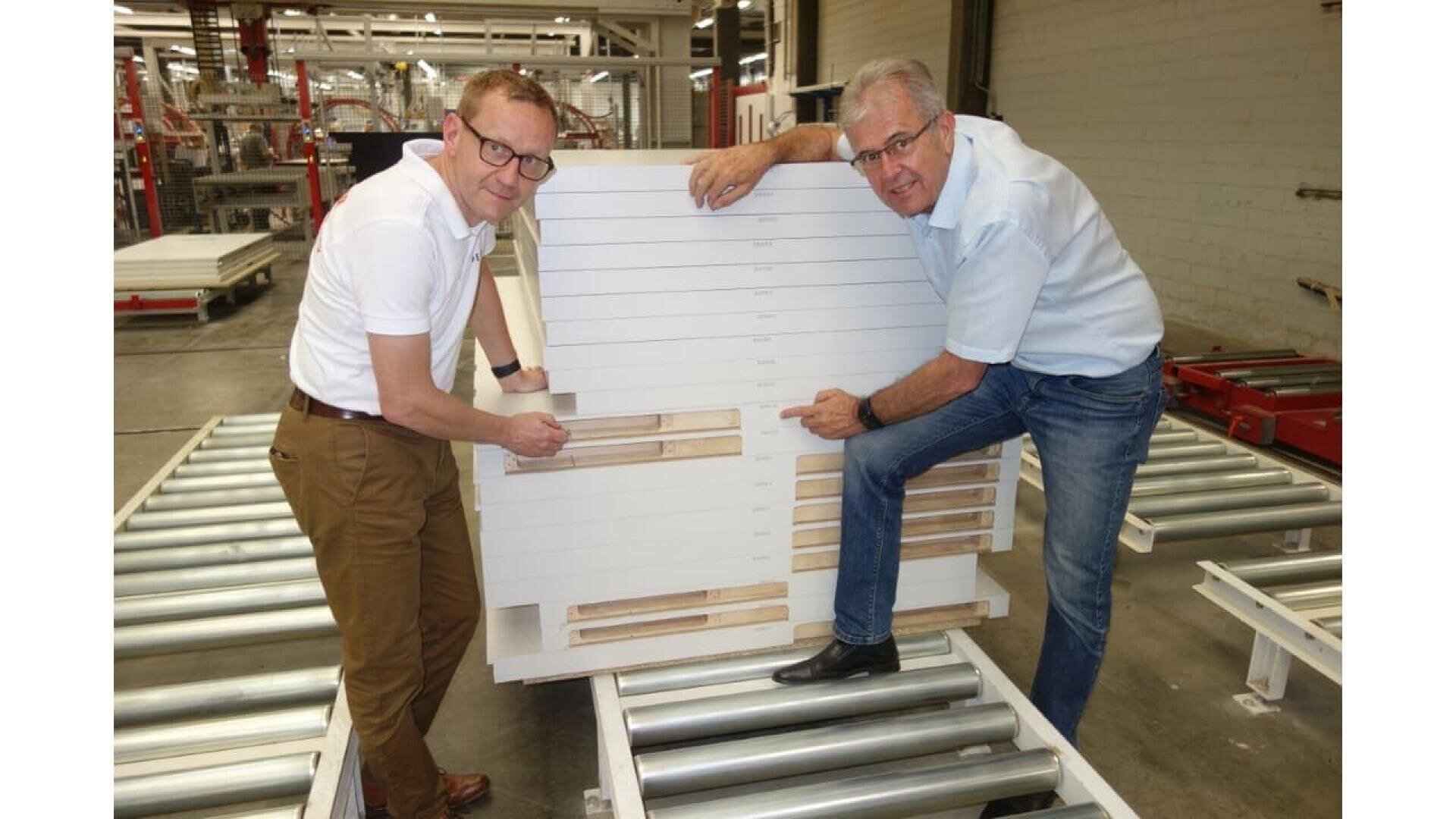 Deux hommes portant des lunettes se tiennent dans une usine, chacun montrant des sections d'un grand panneau blanc à compartiments, placé sur des barres de convoyeurs à rouleaux. Des étagères et des machines sont visibles à l'arrière-plan.