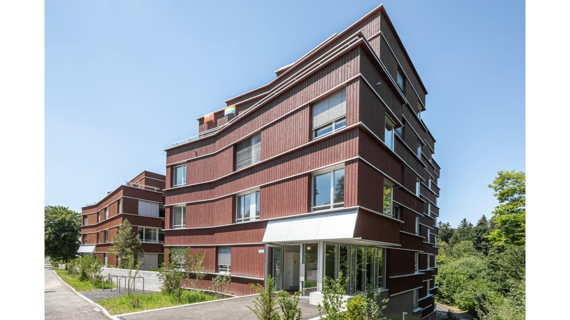 Immeuble moderne de plusieurs étages avec un bardage en bois rouge-brun, de grandes fenêtres et des lignes irrégulières et angulaires. Le bâtiment est entouré de verdure et d'arbres sous un ciel bleu clair.