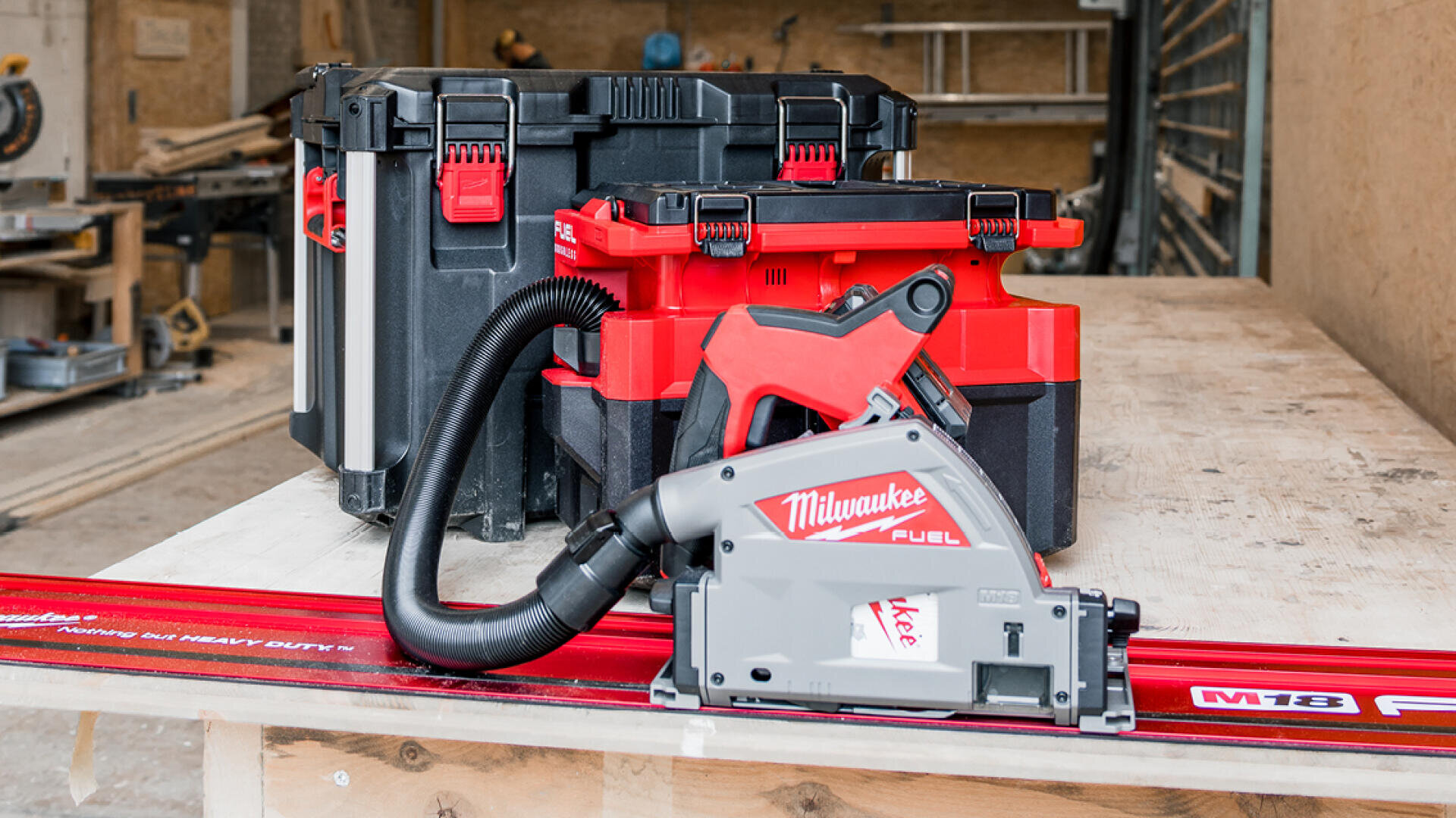 Une scie à chaîne Milwaukee Fuel avec un tuyau d'aspiration est positionnée sur un rail de guidage rouge, devant deux caisses d'outils Milwaukee empilées sur un établi dans un atelier.