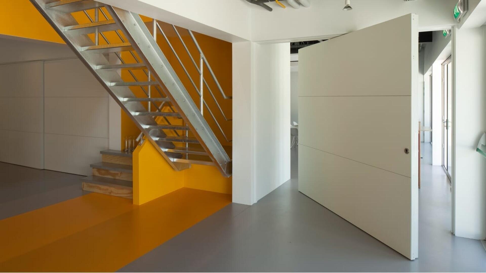 Un intérieur moderne avec un escalier en métal, un mur d'accent jaune vif et une grande porte blanche pivotante partiellement ouverte, révélant une autre pièce au-delà. L'espace présente un design minimaliste aux lignes épurées.