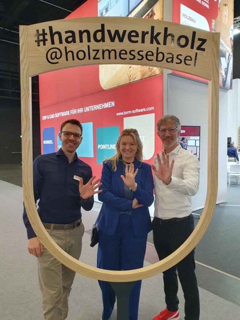 Drei Personen, die lächelnd ihre Hände hochhalten und jeweils fünf Finger zeigen, während sie hinter einem grossen Holzrahmen mit der Aufschrift #handwerkholz @holzmessebasel an einer Indoor-Ausstellung oder Messe posieren.