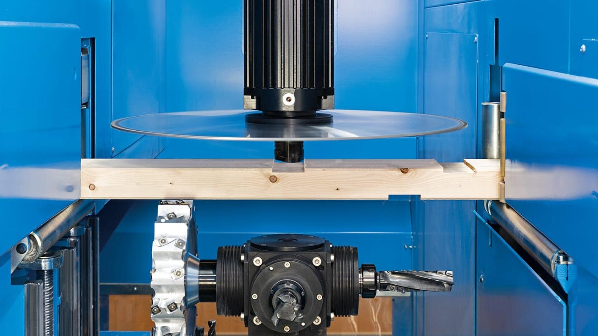 Nahaufnahme einer Industriemaschine mit einem Kreissägeblatt, einem Stück Holz, das bearbeitet wird, und verschiedenen mechanischen Teilen in einem blauen Metallgehäuse.