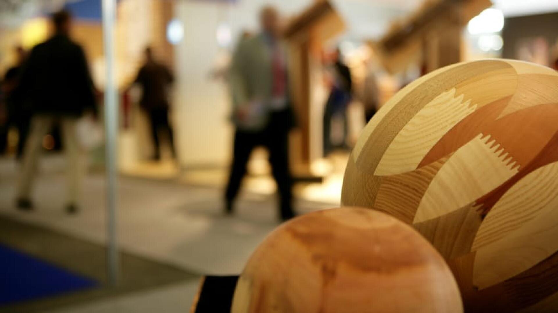 Große Holzkugeln mit geometrischen Mustern im Vordergrund, mit unscharfen Menschen und Ausstellungsstücken in einer hellen Innenumgebung im Hintergrund.