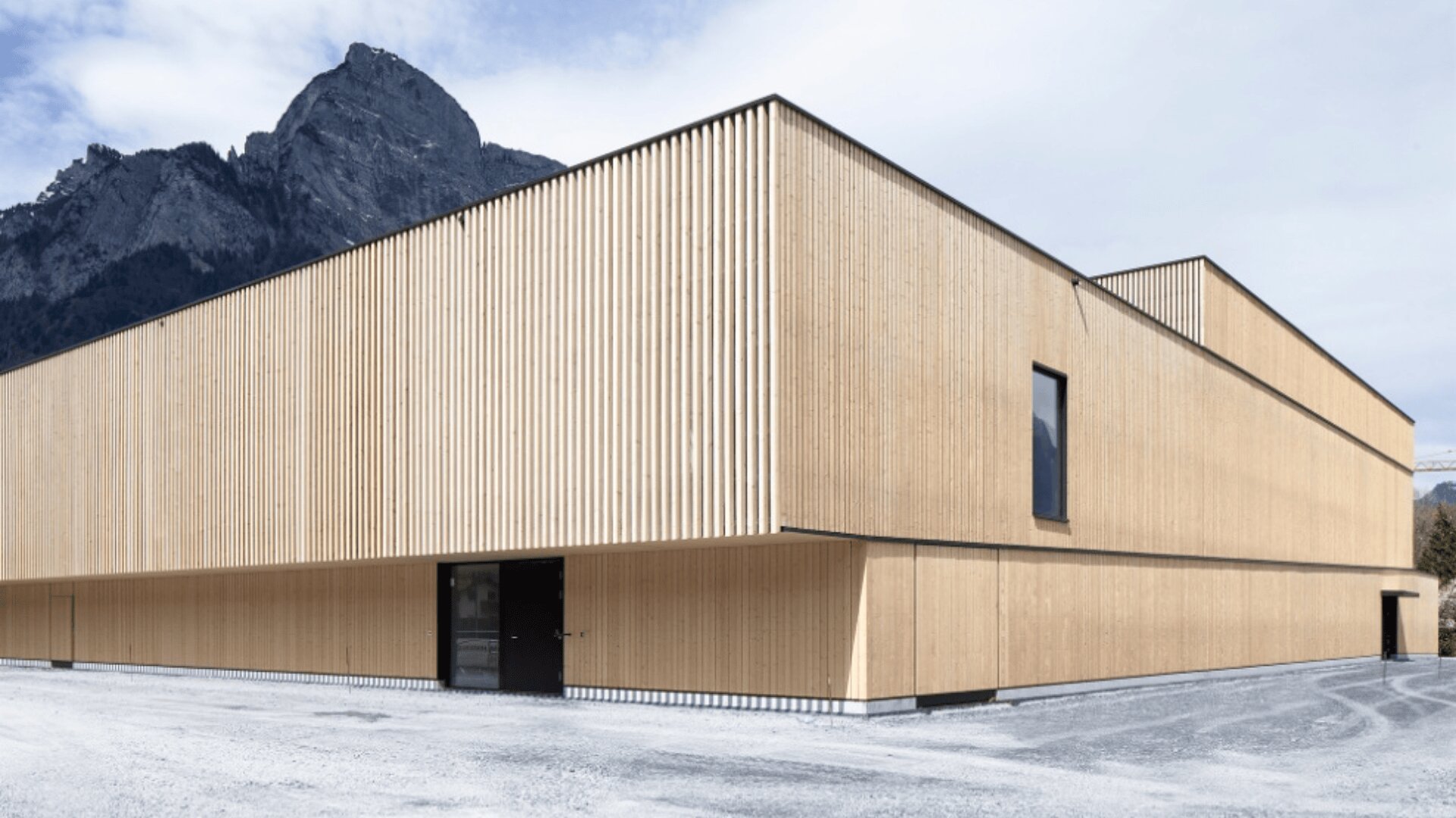 Ein modernes Gebäude mit heller Holzvertäfelung und minimalen Fenstern steht auf einem Kiesgrundstück, mit einem schroffen Berg und einem bewölkten Himmel im Hintergrund.