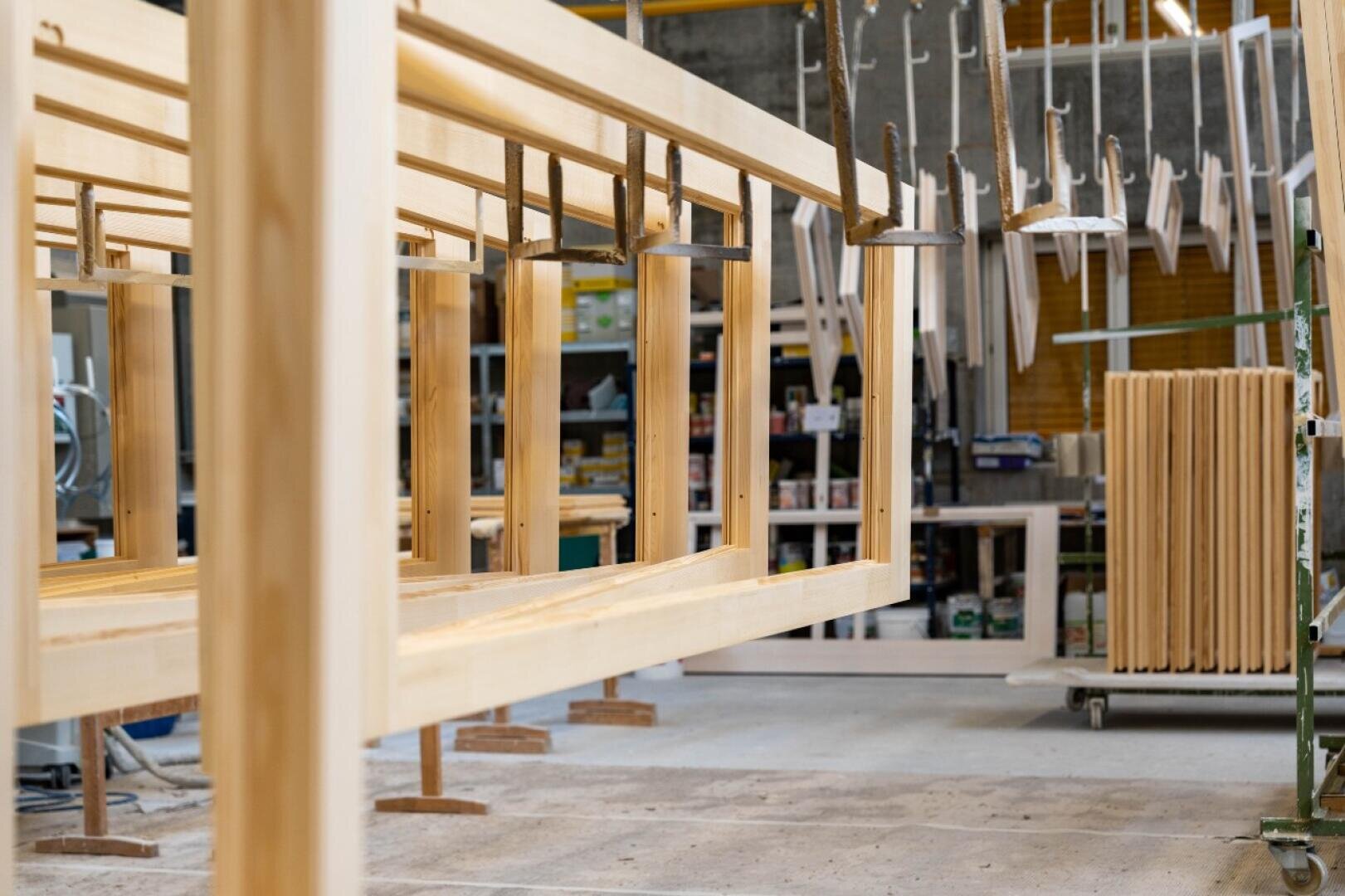 In einer Werkstatt werden hölzerne Möbelrahmen mit Klammern zusammengehalten. Im Hintergrund sind Regale mit Werkzeugen und Materialien sowie andere Holzteile zu sehen. Der Arbeitsbereich ist geordnet und gut beleuchtet.