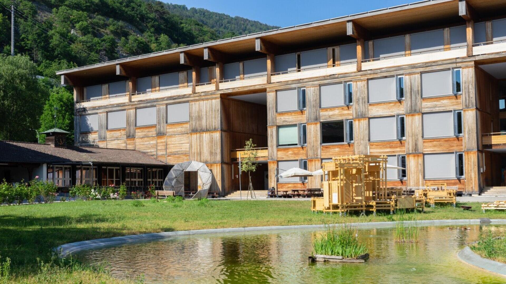 Un bâtiment moderne en bois avec de nombreux stores de fenêtre se trouve derrière un petit étang et une pelouse verte, entourée d'arbres et de collines. Des sièges jaunes et des parasols sont installés dans le jardin.