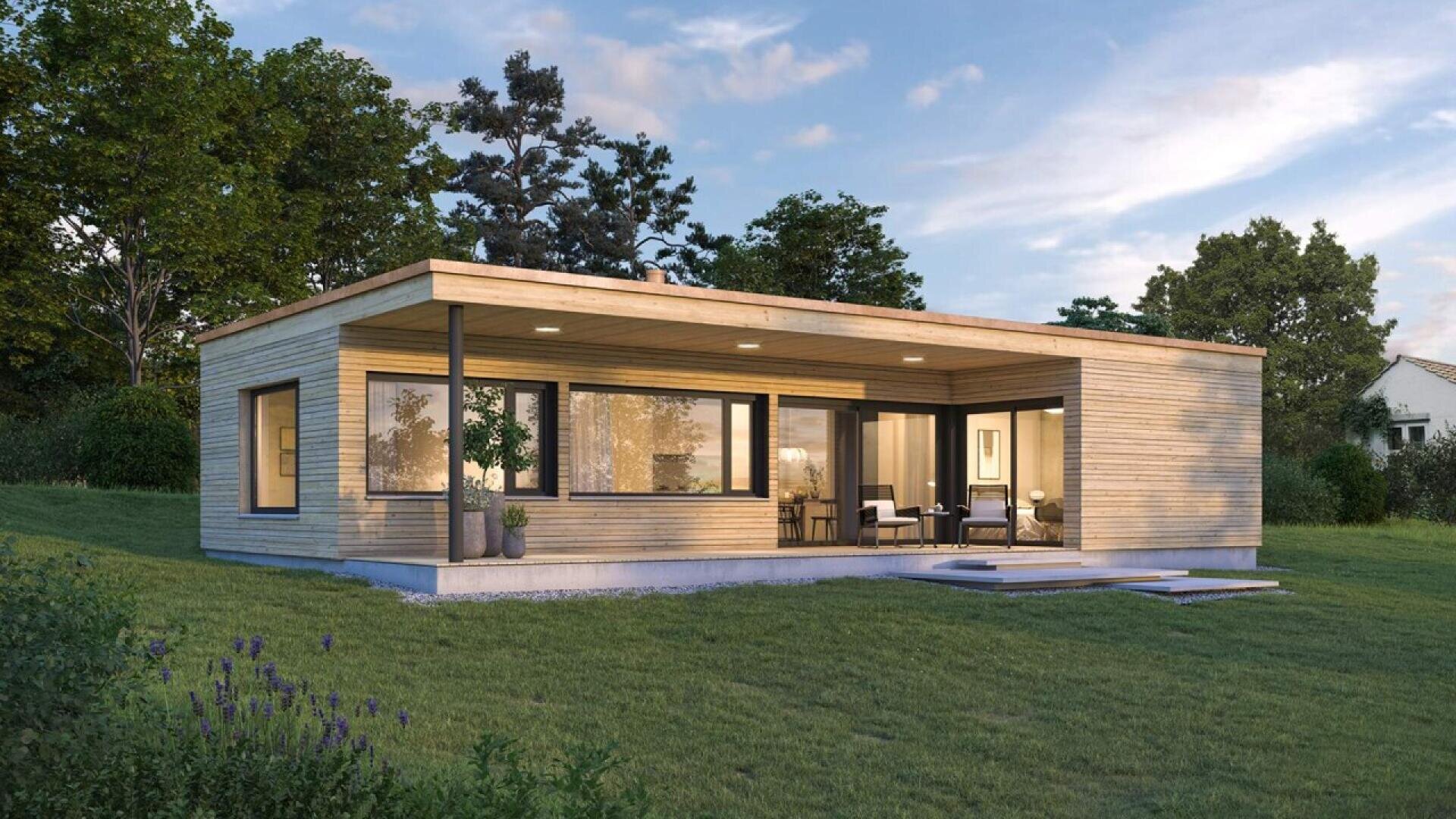 Une maison moderne de plain-pied avec de grandes fenêtres, un bardage en bois et un patio couvert se trouve sur une pelouse entourée d'arbres sous un ciel partiellement nuageux.