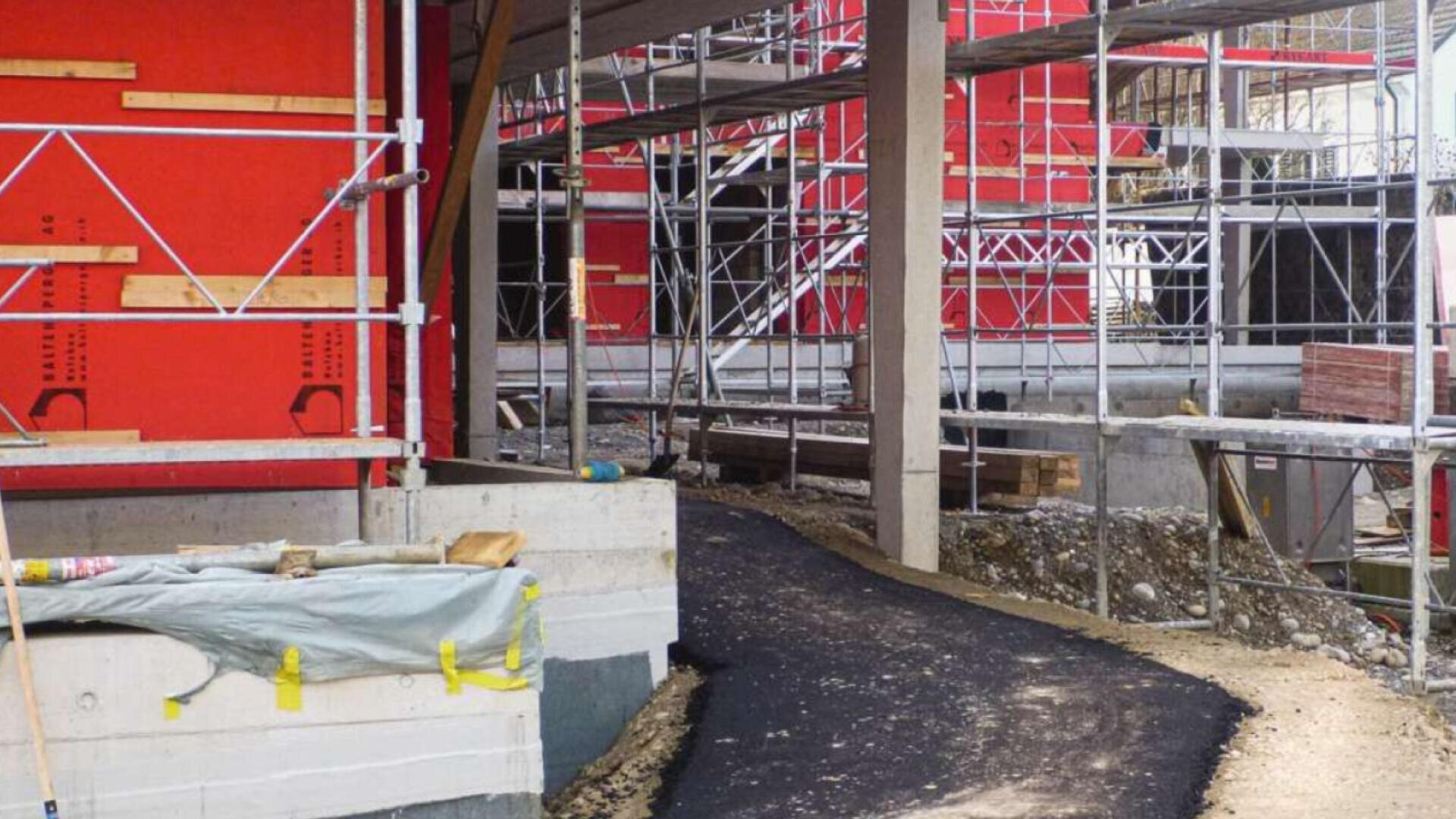 Eine Baustelle mit roten Gerüsten, Betonwänden und einem neu gepflasterten Weg aus schwarzem Asphalt, der sich zwischen den Gebäuden schlängelt. An den Rändern sind Baumaterialien und Werkzeuge zu sehen.