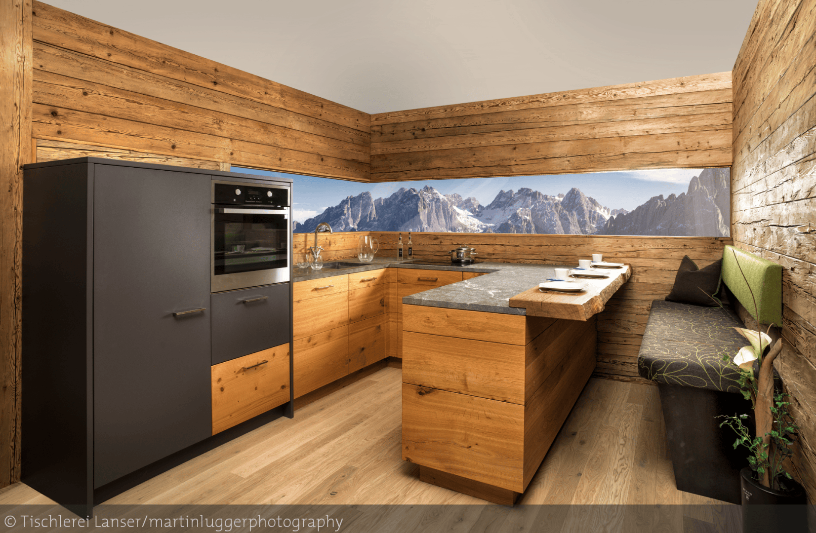 Eine moderne Küchenzeile mit holzgetäfelten Wänden, schlichten schwarzen und hölzernen Schränken, einem eingebauten Ofen und einer Arbeitsplatte aus Stein. Durch ein Fenster hat man einen Blick auf die Berge, und eine Eckbank mit grünen Kissen bietet Sitzgelegenheiten.