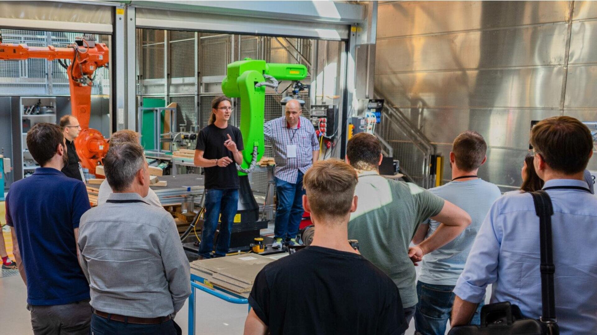 Un groupe de personnes écoute deux présentateurs dans un atelier industriel équipé de bras robotisés et de machines. Le cadre semble moderne et high-tech, avec un éclairage vif et des surfaces métalliques.