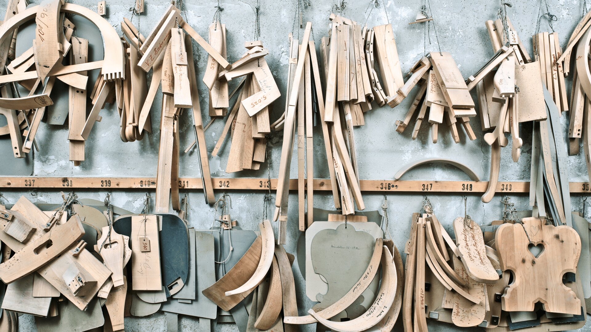 Verschiedene Holzschablonen und Teile für Musikinstrumente wie Geigen und Celli hängen an Haken an einer Betonwand in einer Werkstatt. Jedes Teil ist beschriftet und nach Form und Größe geordnet.