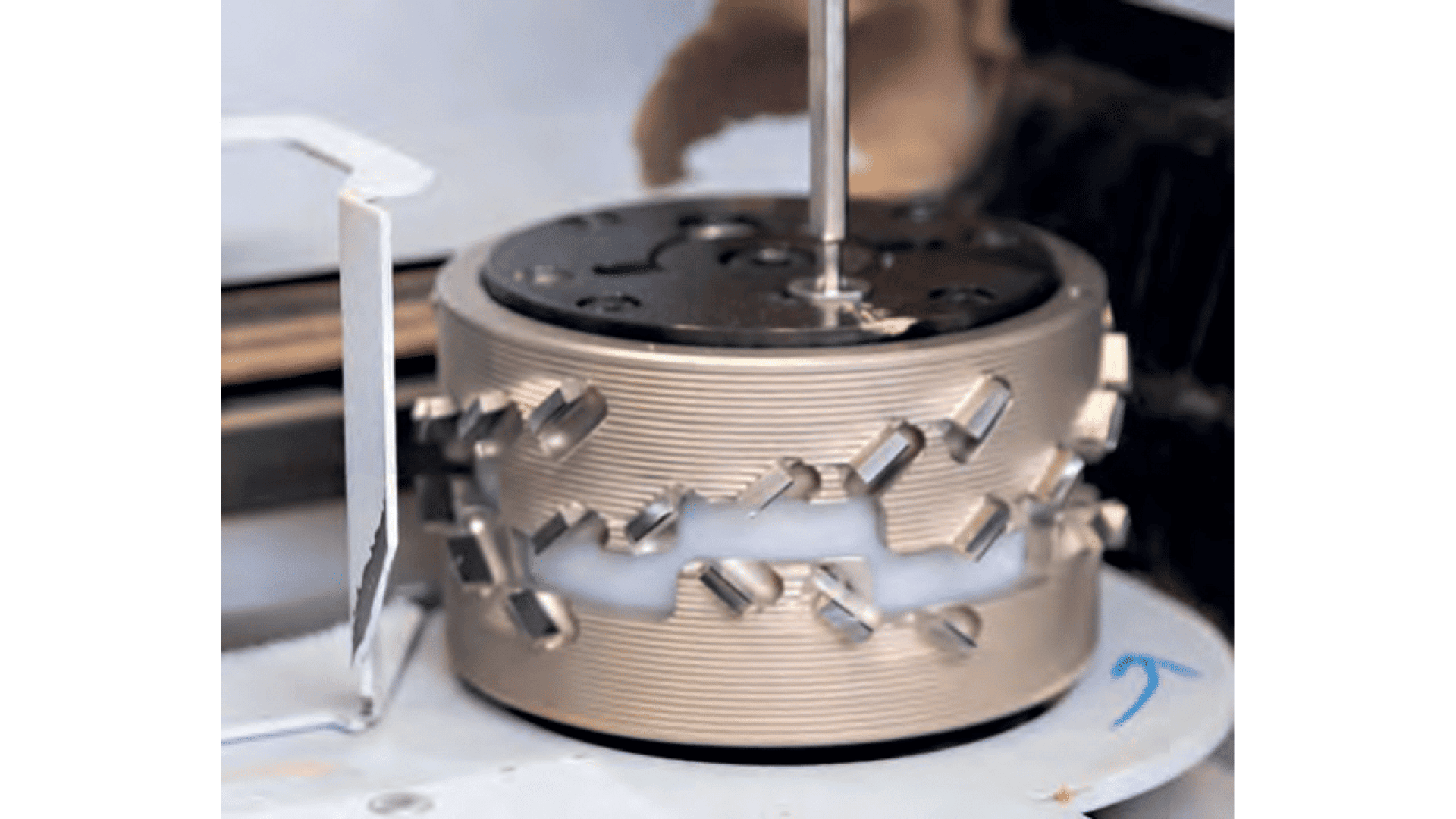 Gros plan d'un objet métallique cylindrique avec des découpes géométriques, monté verticalement sur une machine rotative. Une tige part du centre supérieur et une flèche bleue est marquée sur la base pour indiquer le sens de rotation.