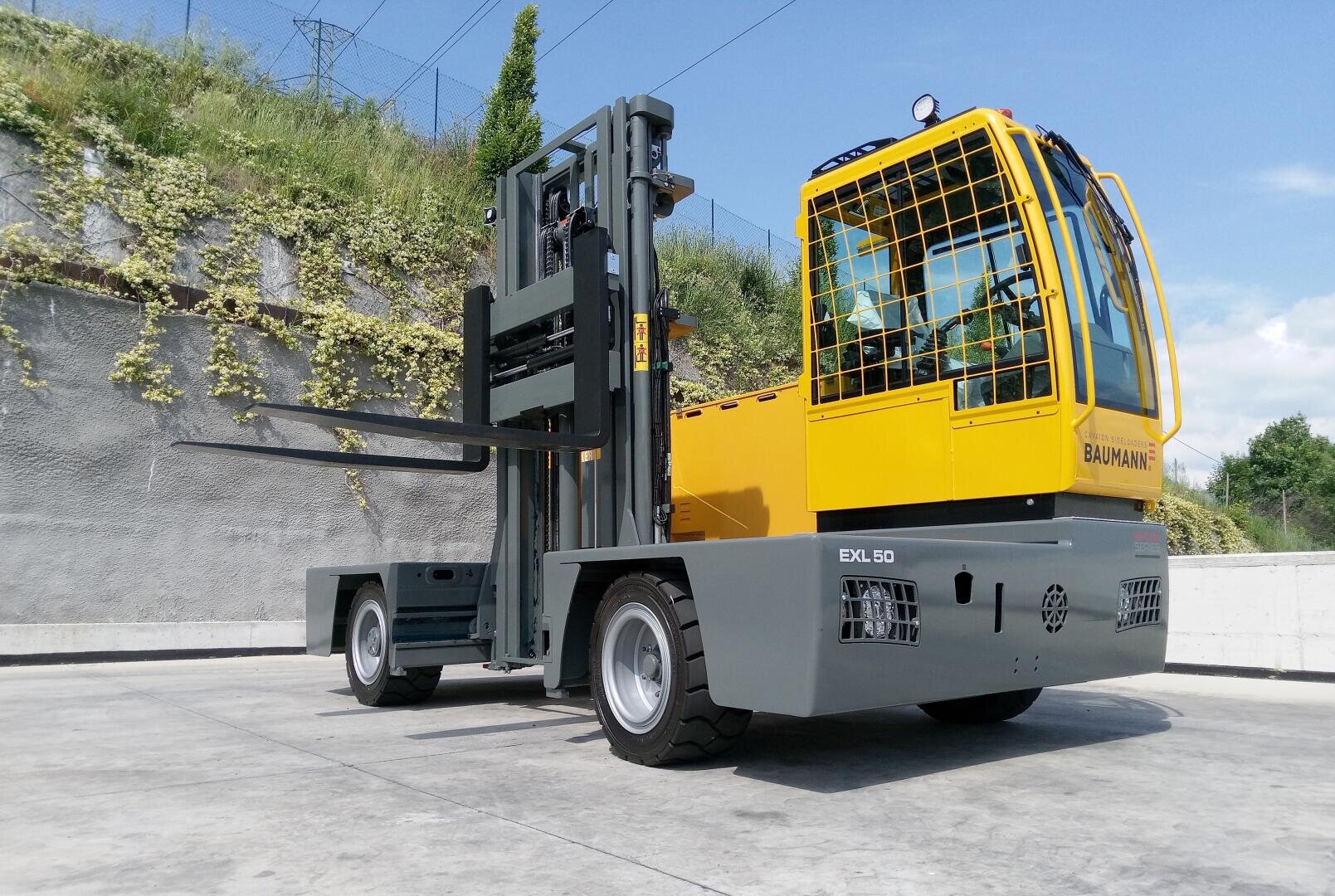 Ein gelber und grauer Baumann EXL 50-Seitenstapler ist auf einer Betonfläche in der Nähe einer Mauer mit grünen Pflanzen und einem blauen Himmel im Hintergrund abgestellt.