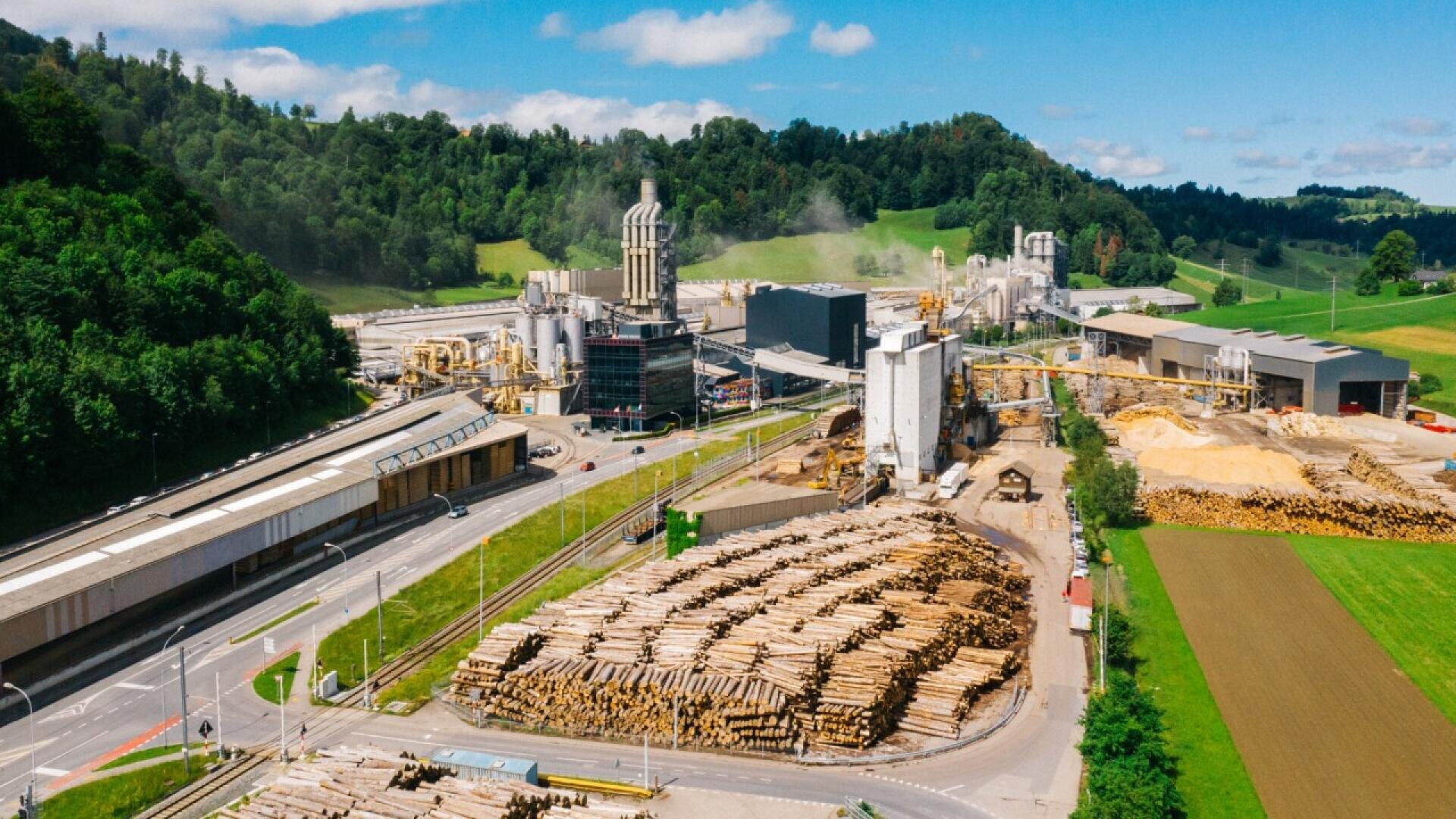 Vue aérienne d'une grande scierie avec des piles de grumes, plusieurs bâtiments industriels, de la fumée s'élevant des cheminées, et des collines vertes et des arbres environnants sous un ciel bleu. Des routes et des champs bordent l'installation.